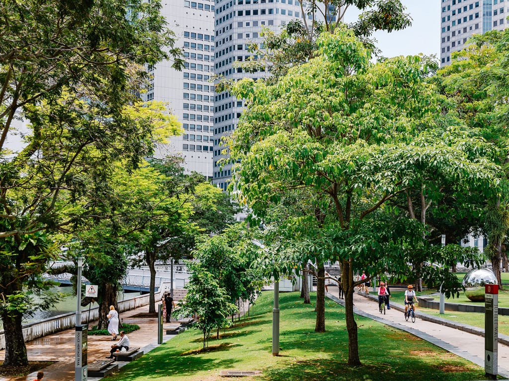 A tree-lined urban park with cyclists and pedestrians on paths, green lawns, and tall office towers in the background.