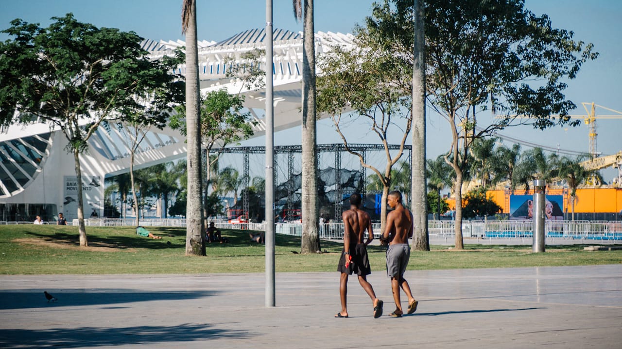 Two shirtless men walk on a paved plaza with palm trees and a white modern building in the background.