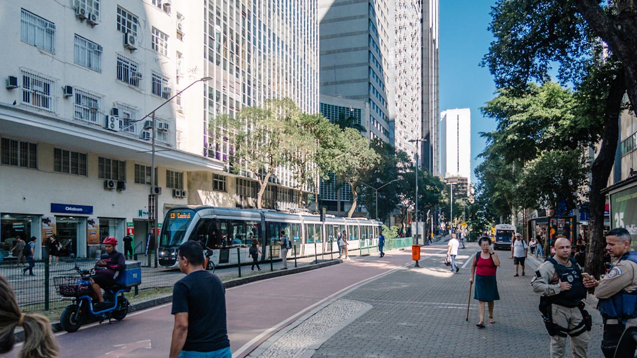 Busy urban street with a modern tram, pedestrians, tall buildings, trees, and two security officers on the right sidewalk.