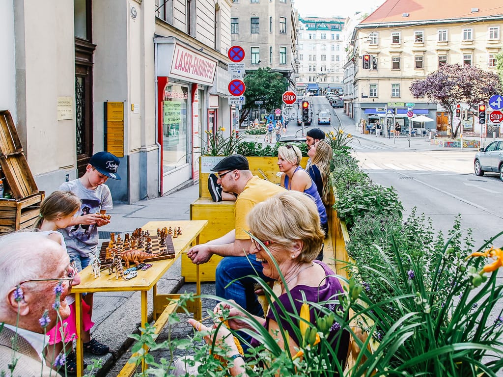 People sitting on yellow outdoor benches near a chess board on a city sidewalk with plants and street signs.