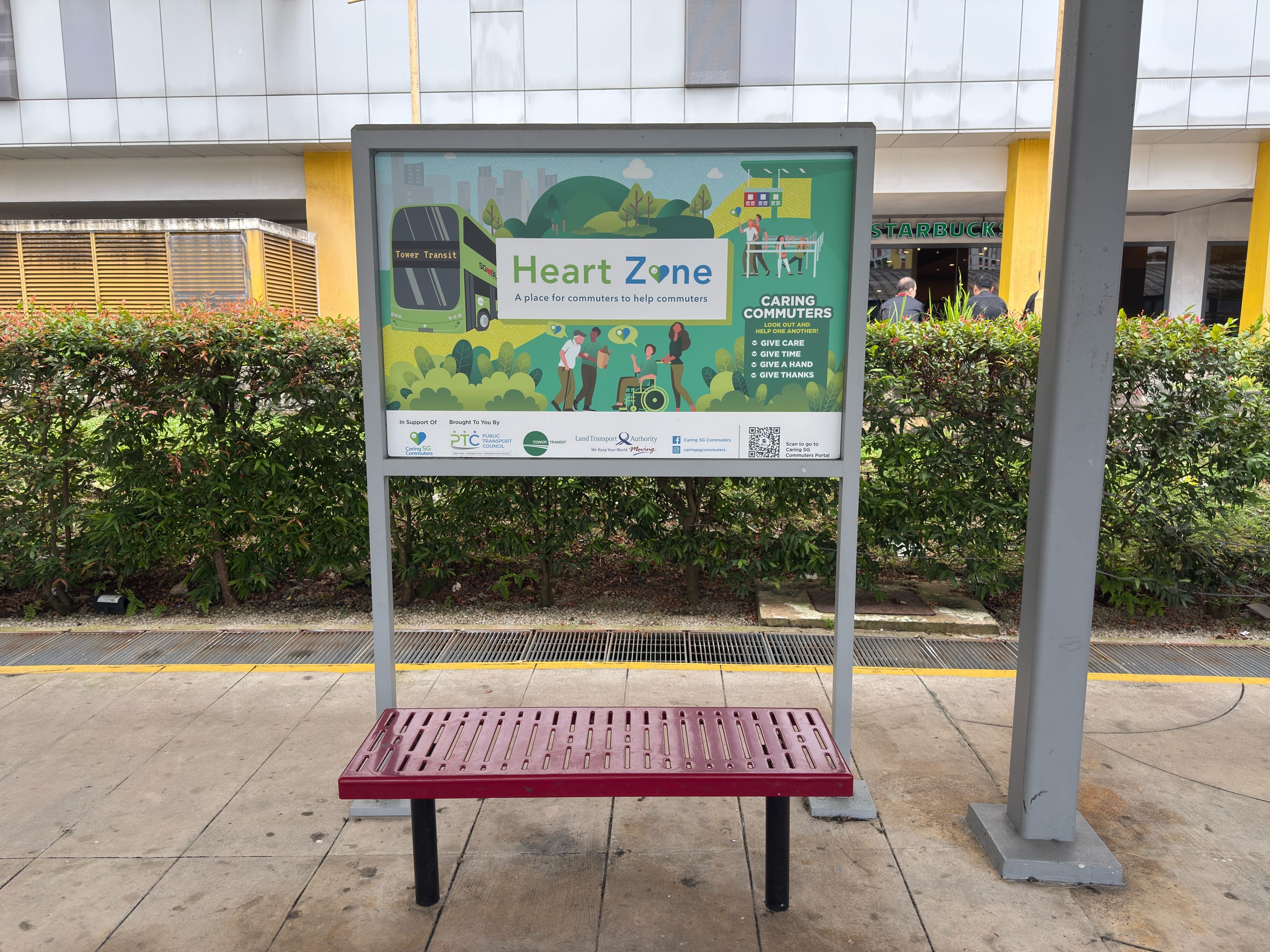Heart Zone signboard outside a bus interchange promoting “Caring Commuters” with benches below the sign.