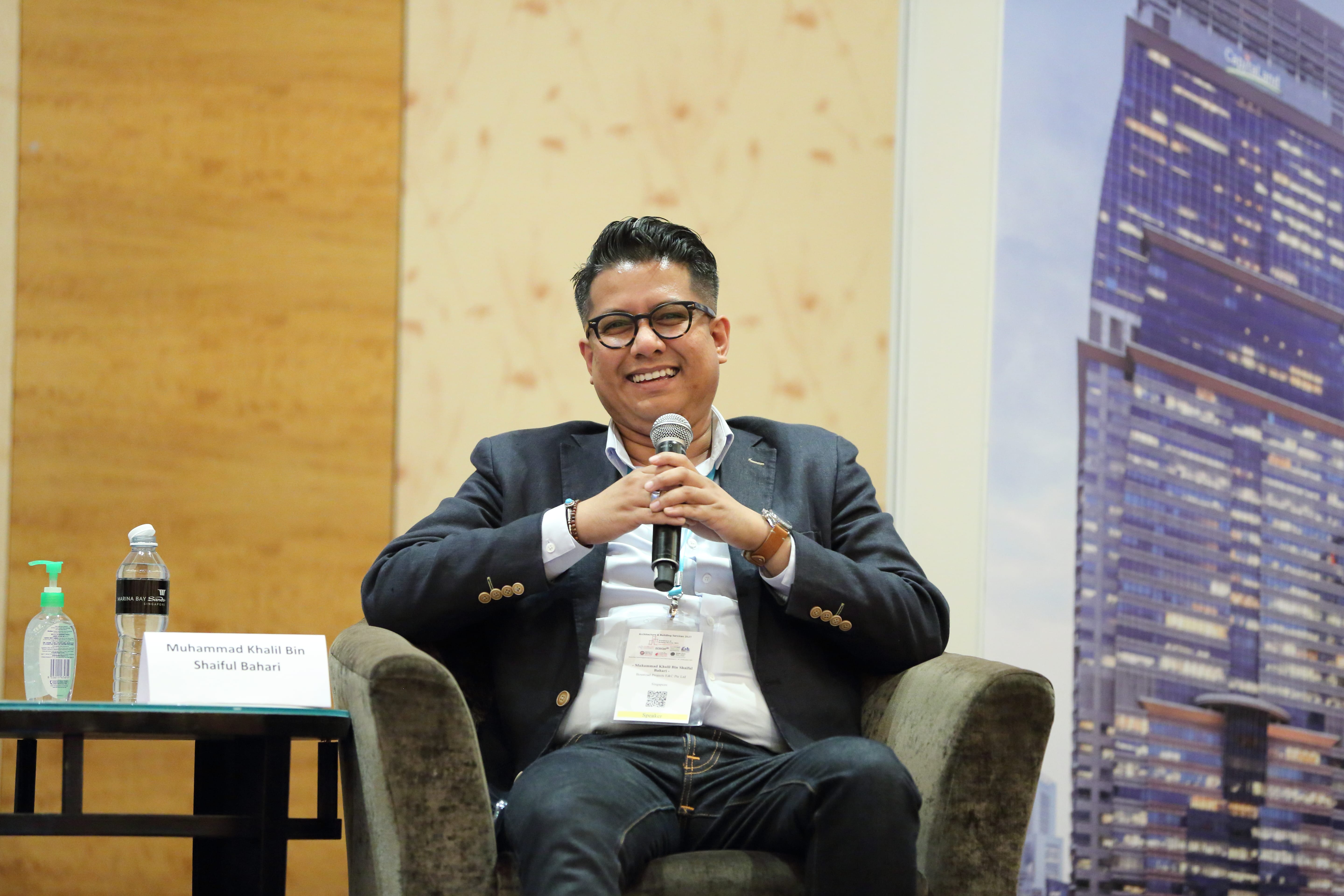 Man with glasses in a blazer sits in a chair, holding a microphone.
