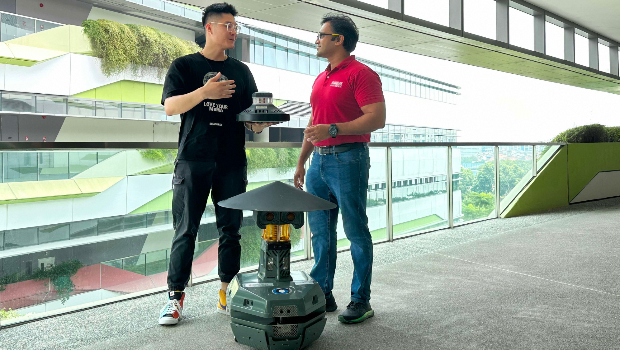 Two men and a gray security robot with yellow lights indoors. One man holds a sensor.