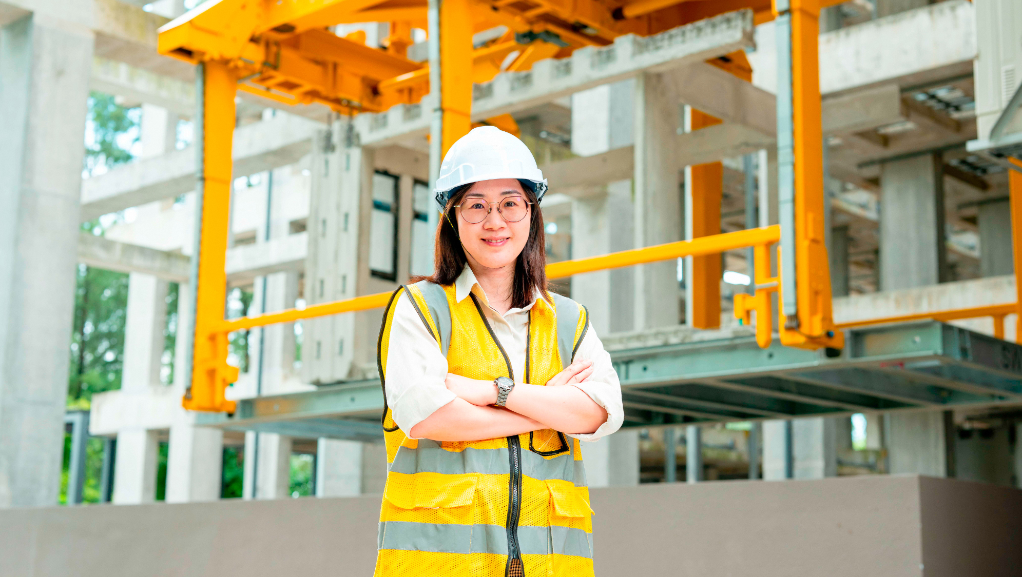 Person in hard hat, safety vest, arms crossed, posing in front of construction site.