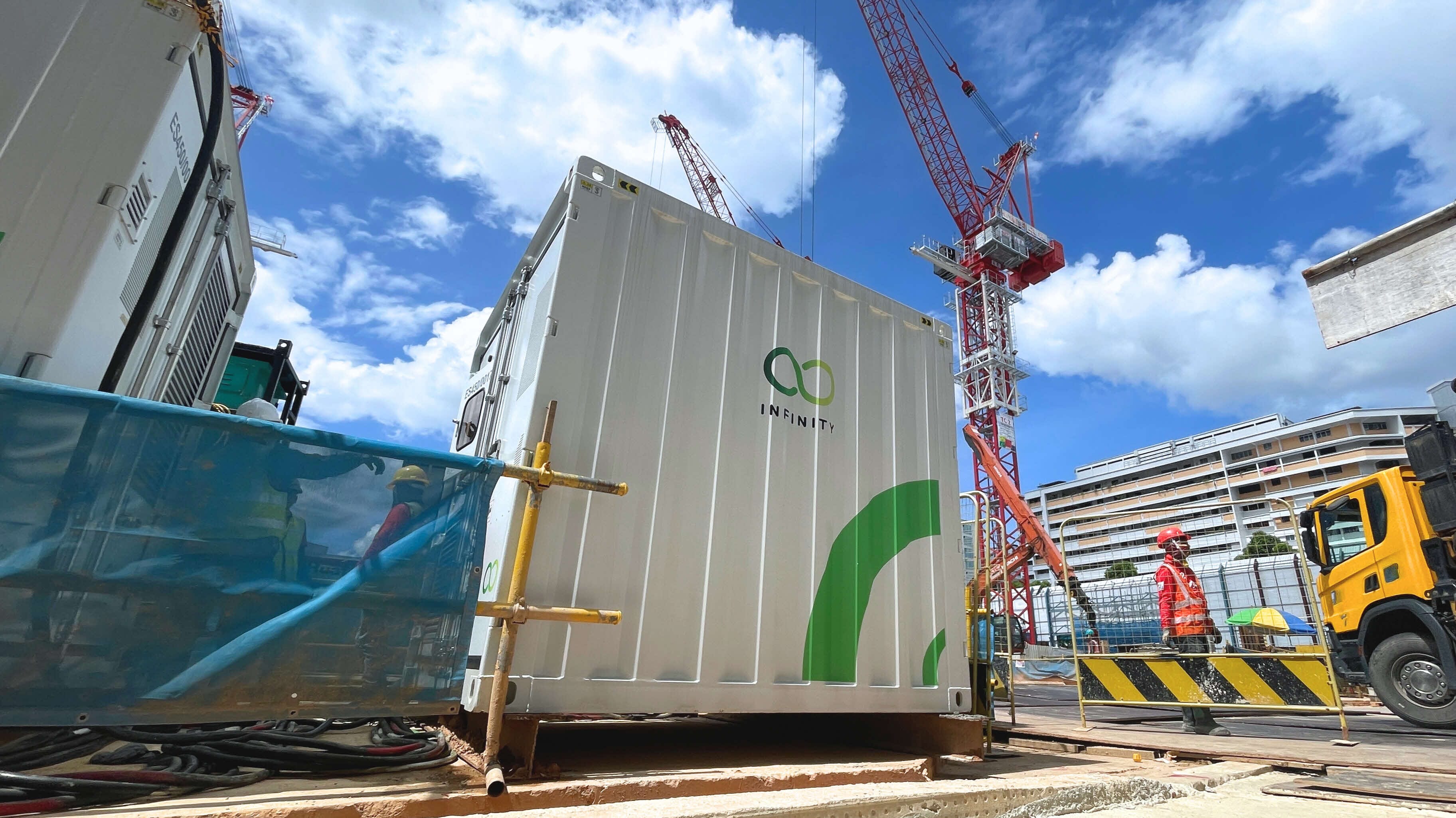 Construction site with white Infinity container, cranes, and workers in vests near a yellow truck.