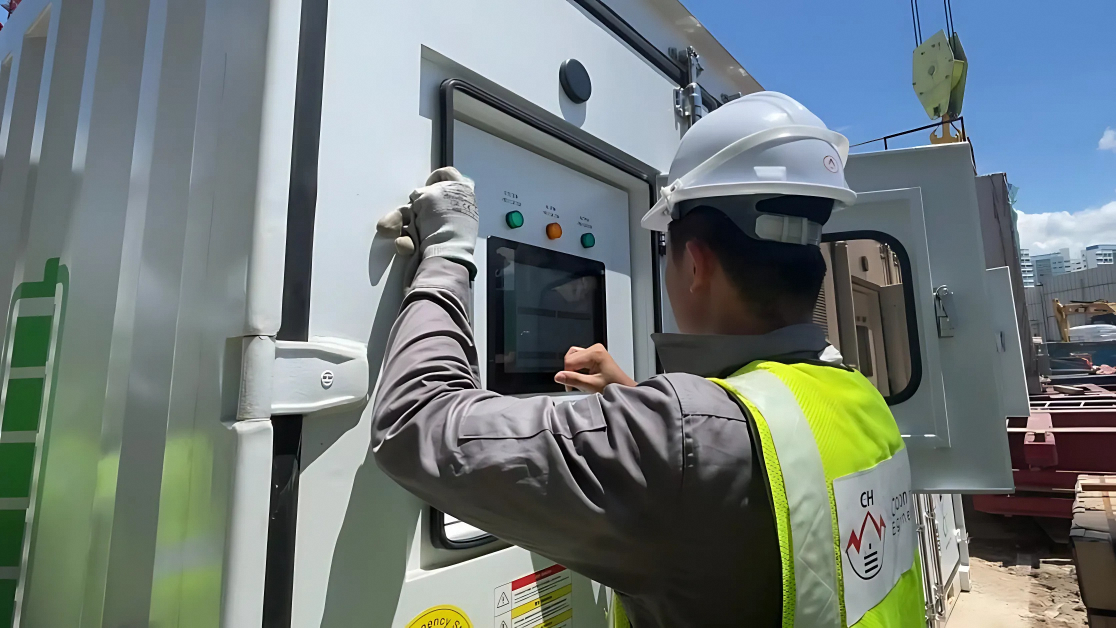 Technician in hard hat adjusts controls on white electrical box with green and orange buttons at a construction site.