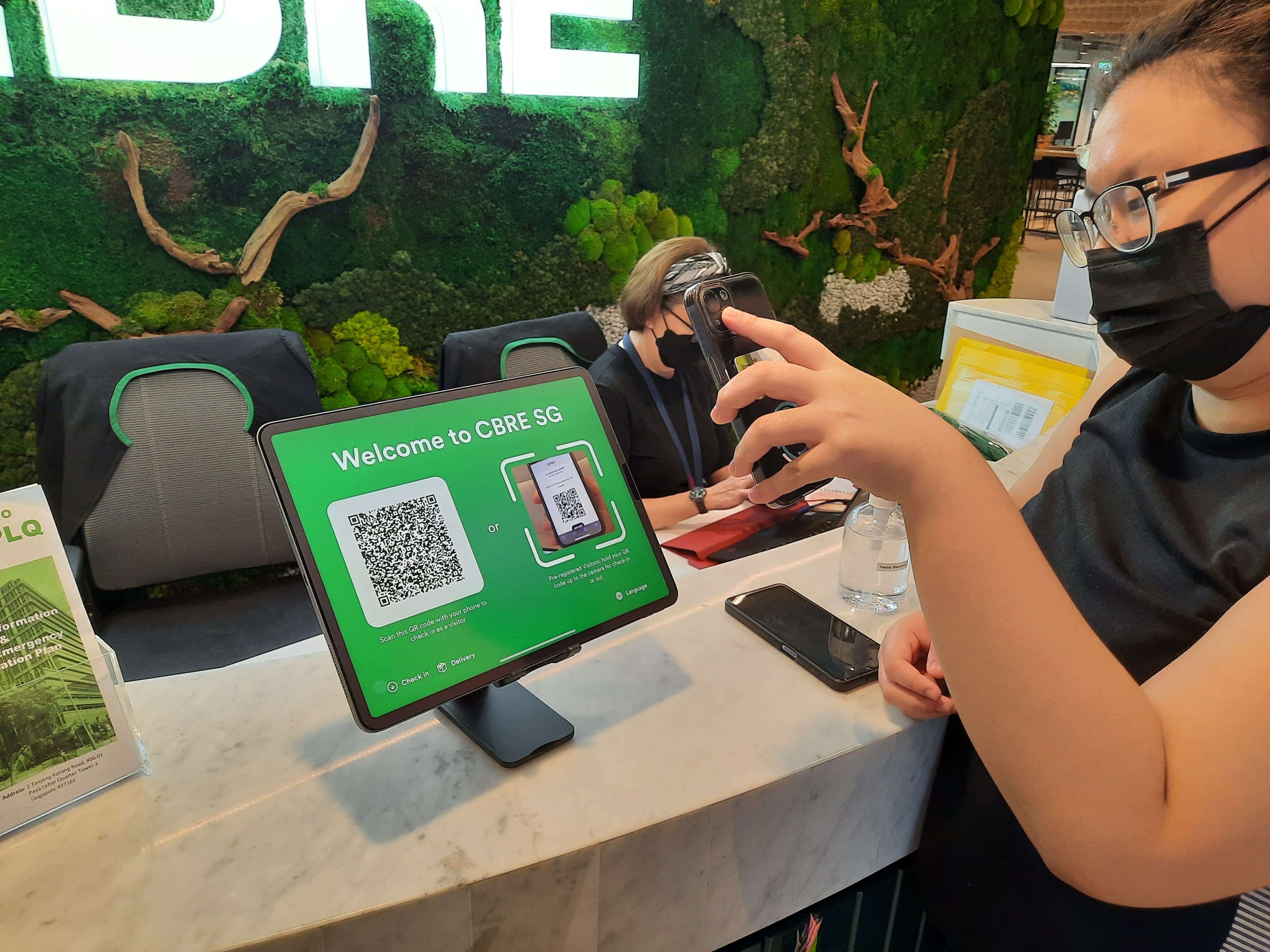 Person scans QR code on a "Welcome to CBRE SG" tablet with phone. Moss wall background.