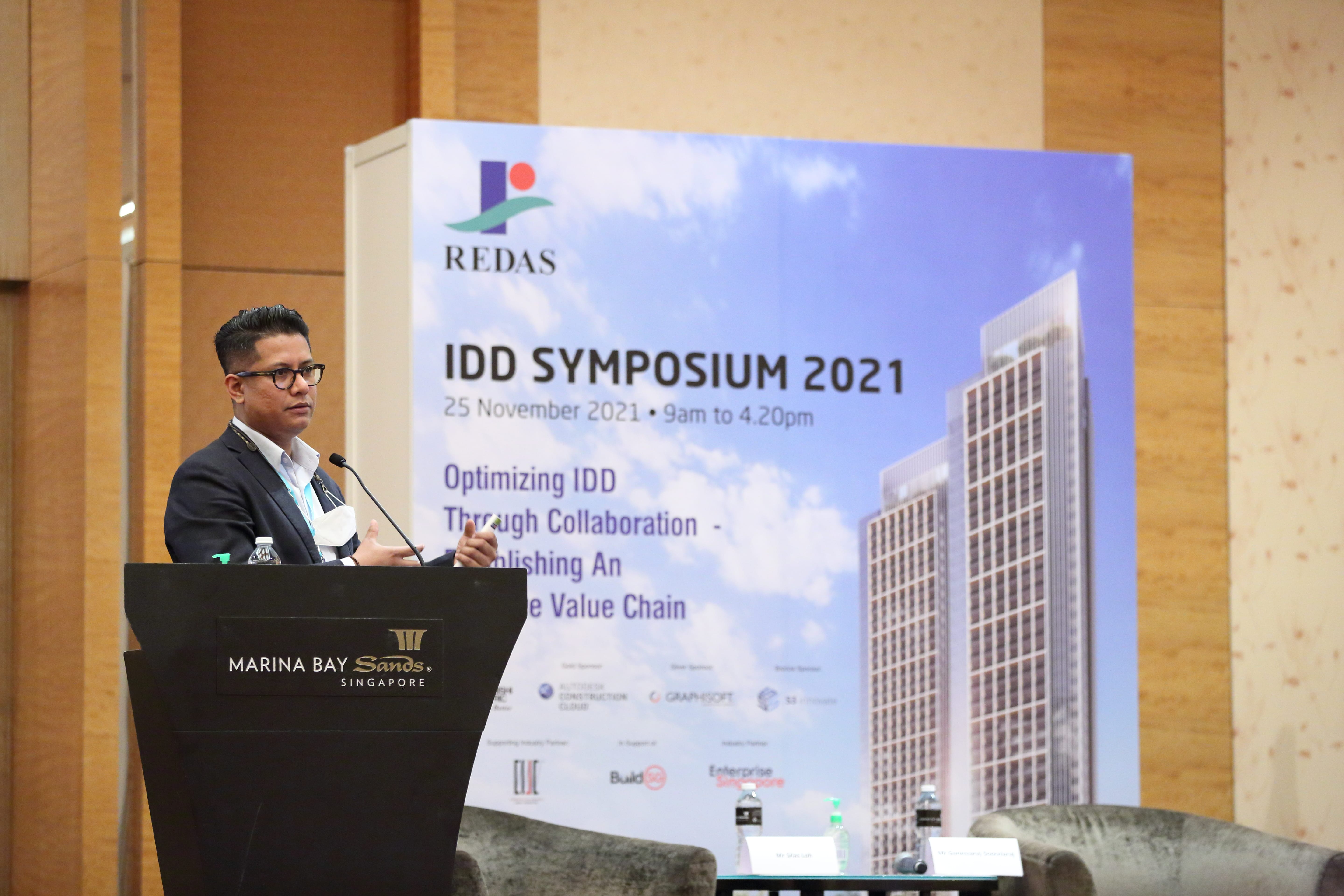 Man speaking at Marina Bay Sands event, "IDD Symposium 2021", with building backdrop.