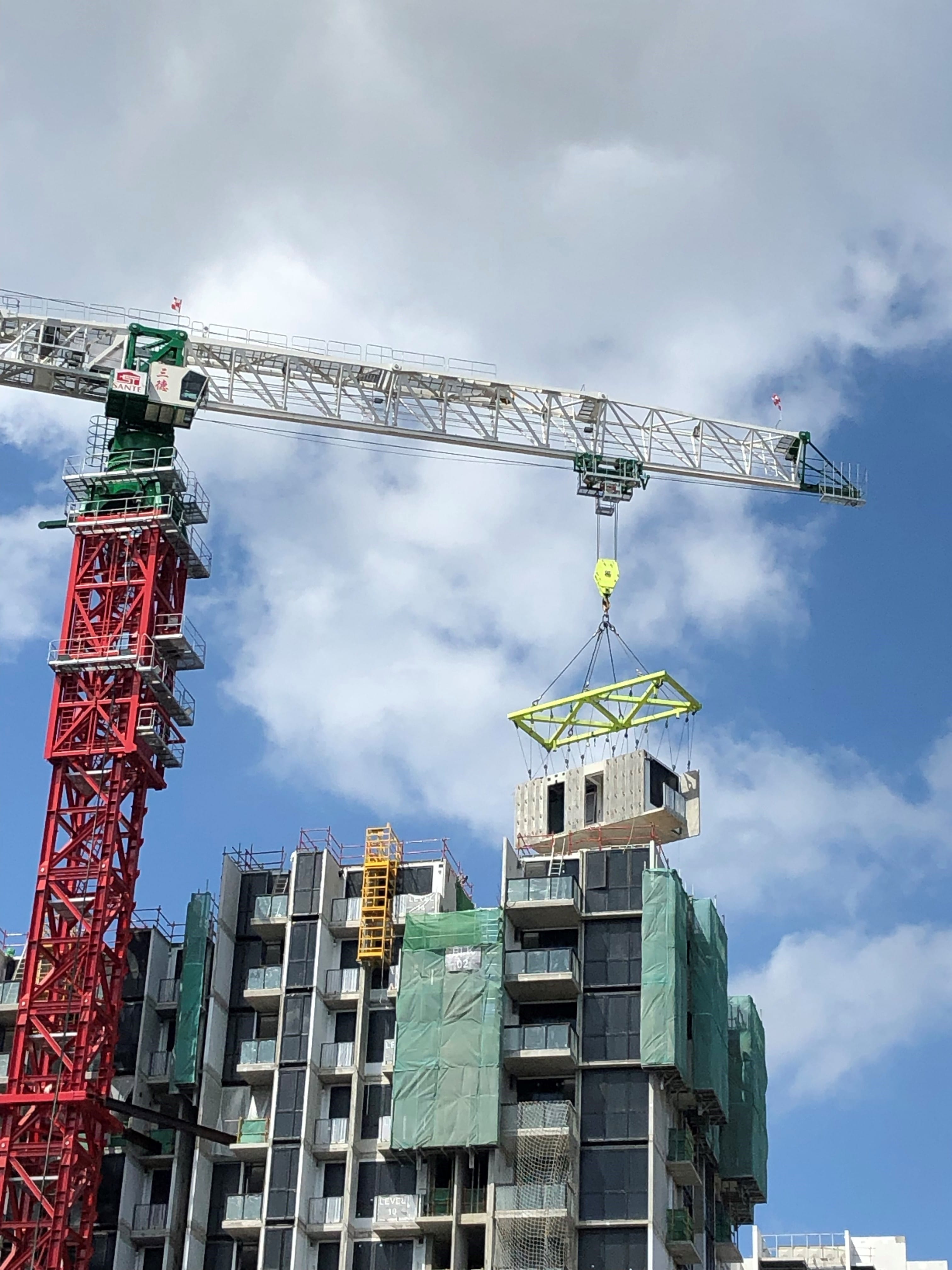 Red and white crane lifting a building section in front of an apartment building under construction.