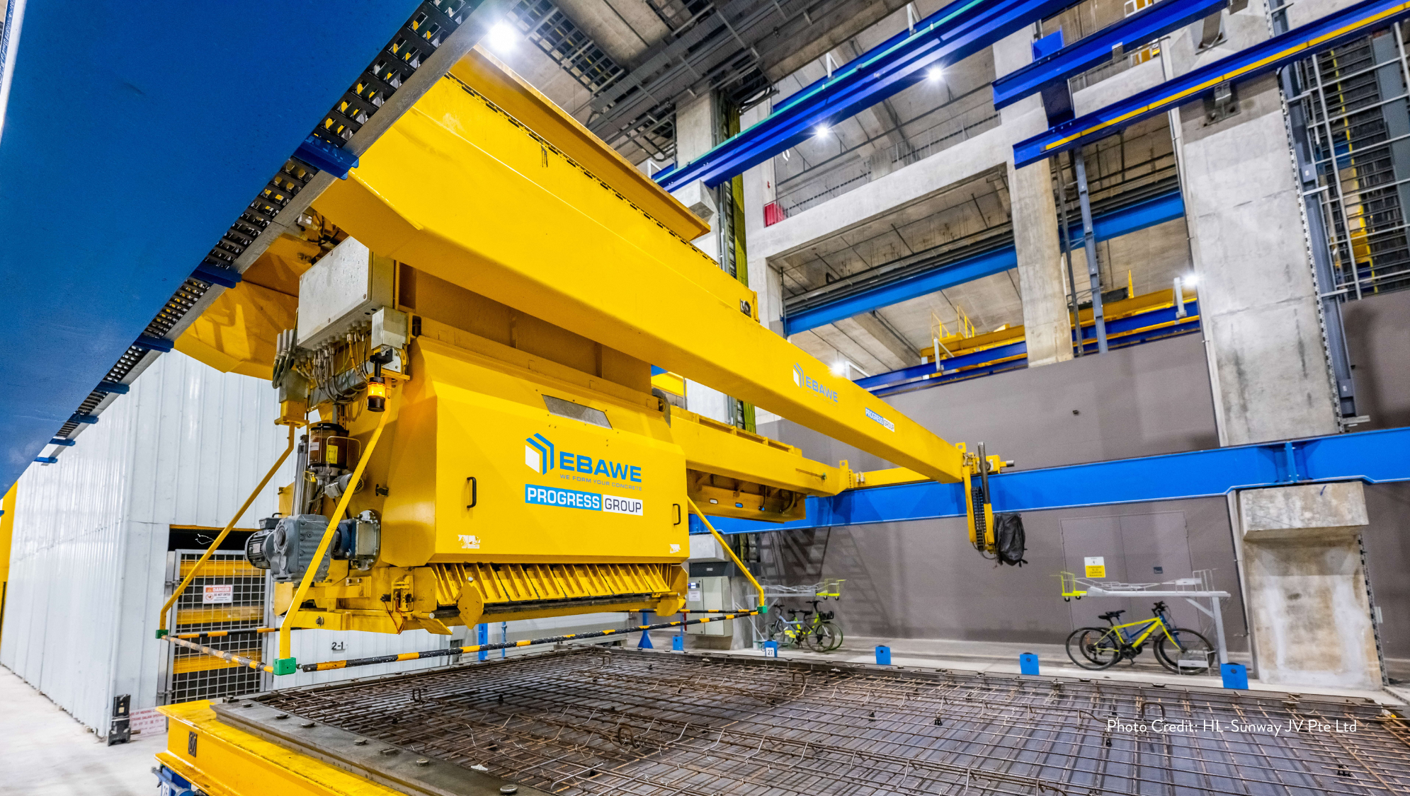 EBAWE Progress Group yellow rebar machine over wire grid in industrial setting, with two parked bicycles nearby.