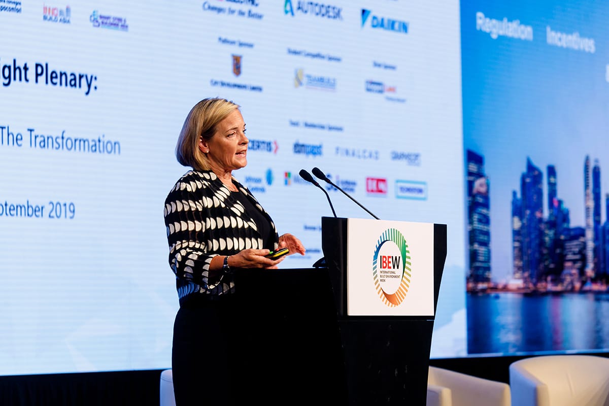 Woman in patterned jacket at lectern with IBEW logo, city skyline on screen behind.