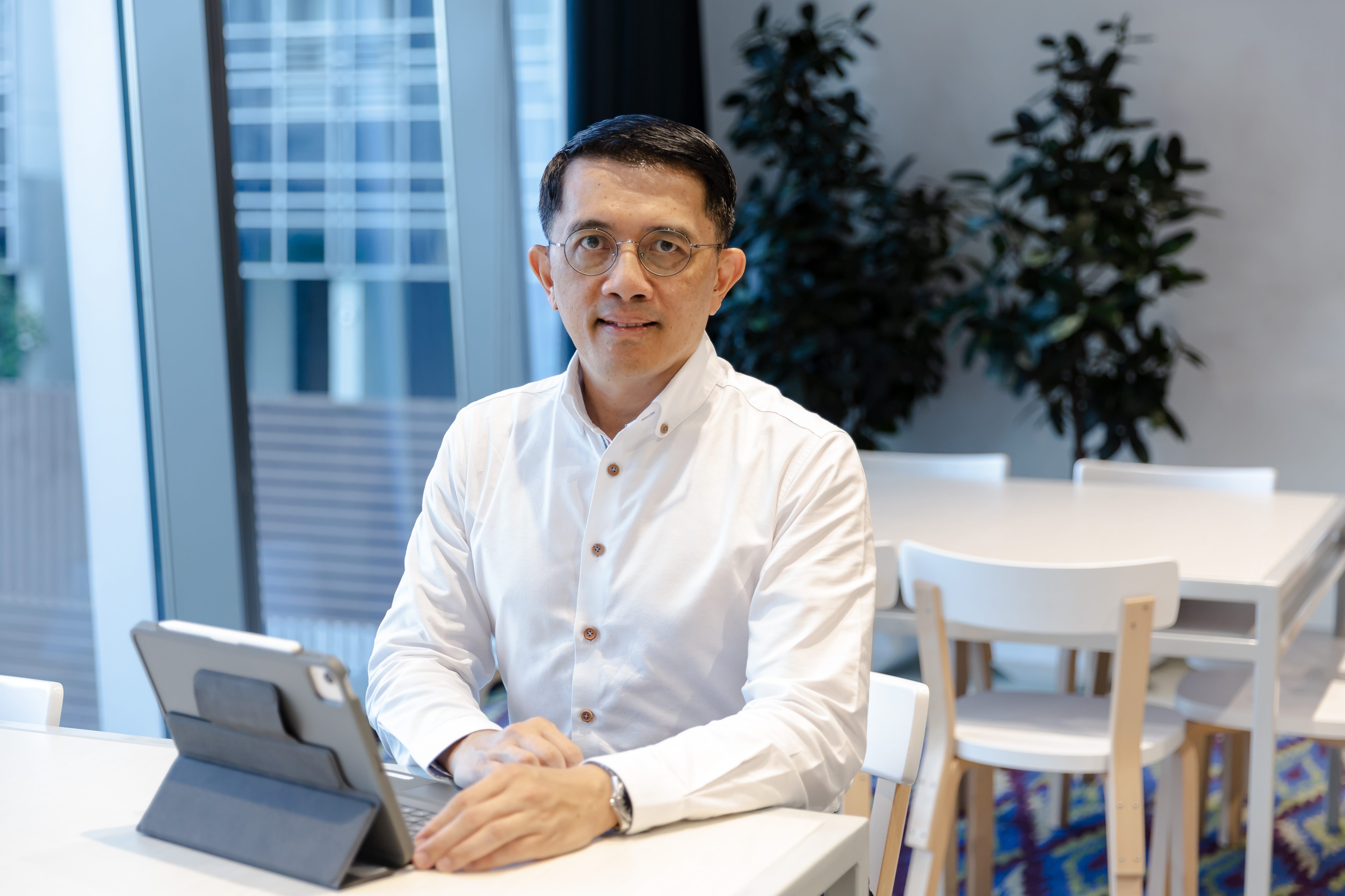 Person with glasses, in a white shirt, sits at a table with a tablet. Modern building in background.