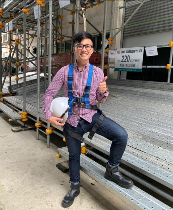 Person in safety harness giving thumbs up, standing on scaffolding and holding hardhat.
