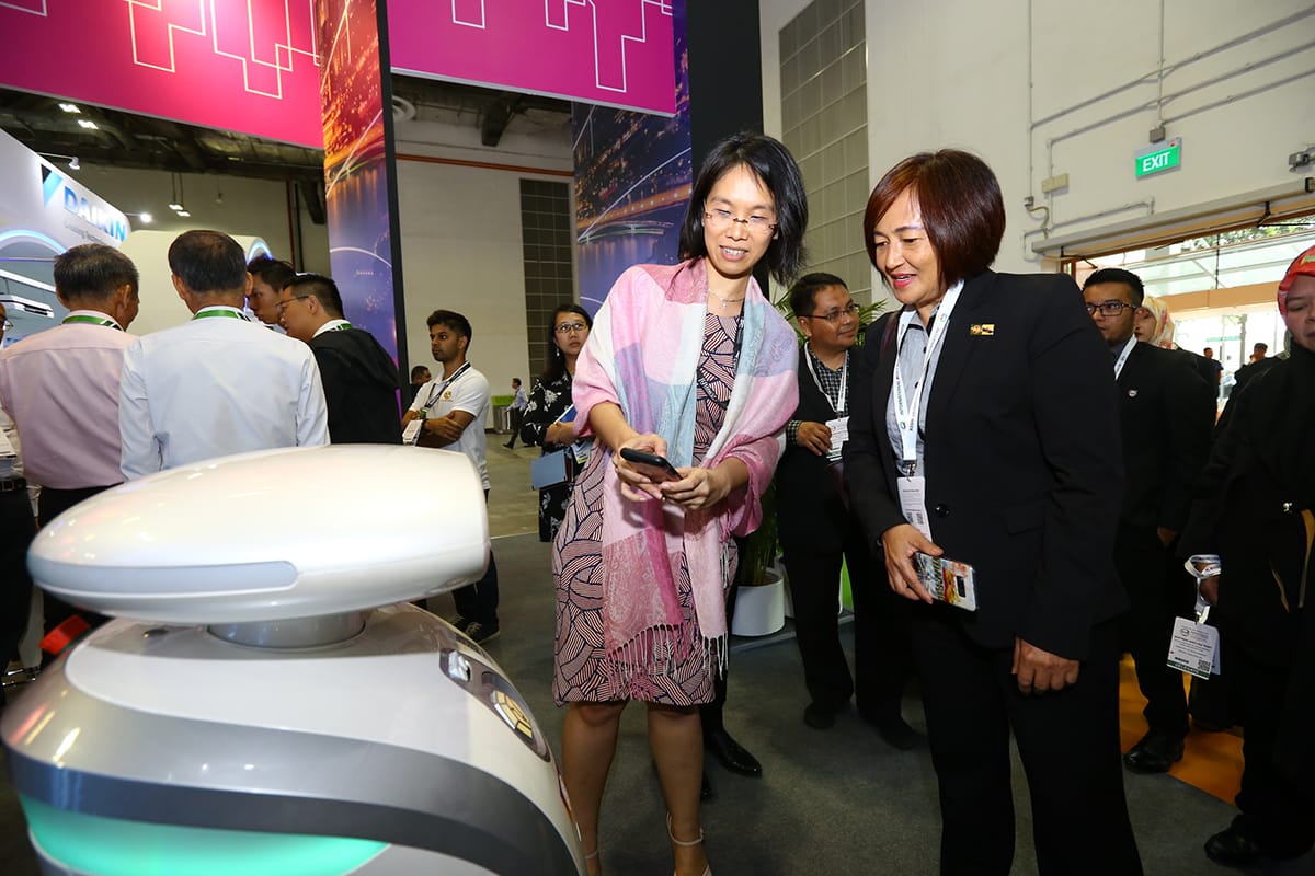 At a trade show, two women look at a white robot; other people are in the background.
