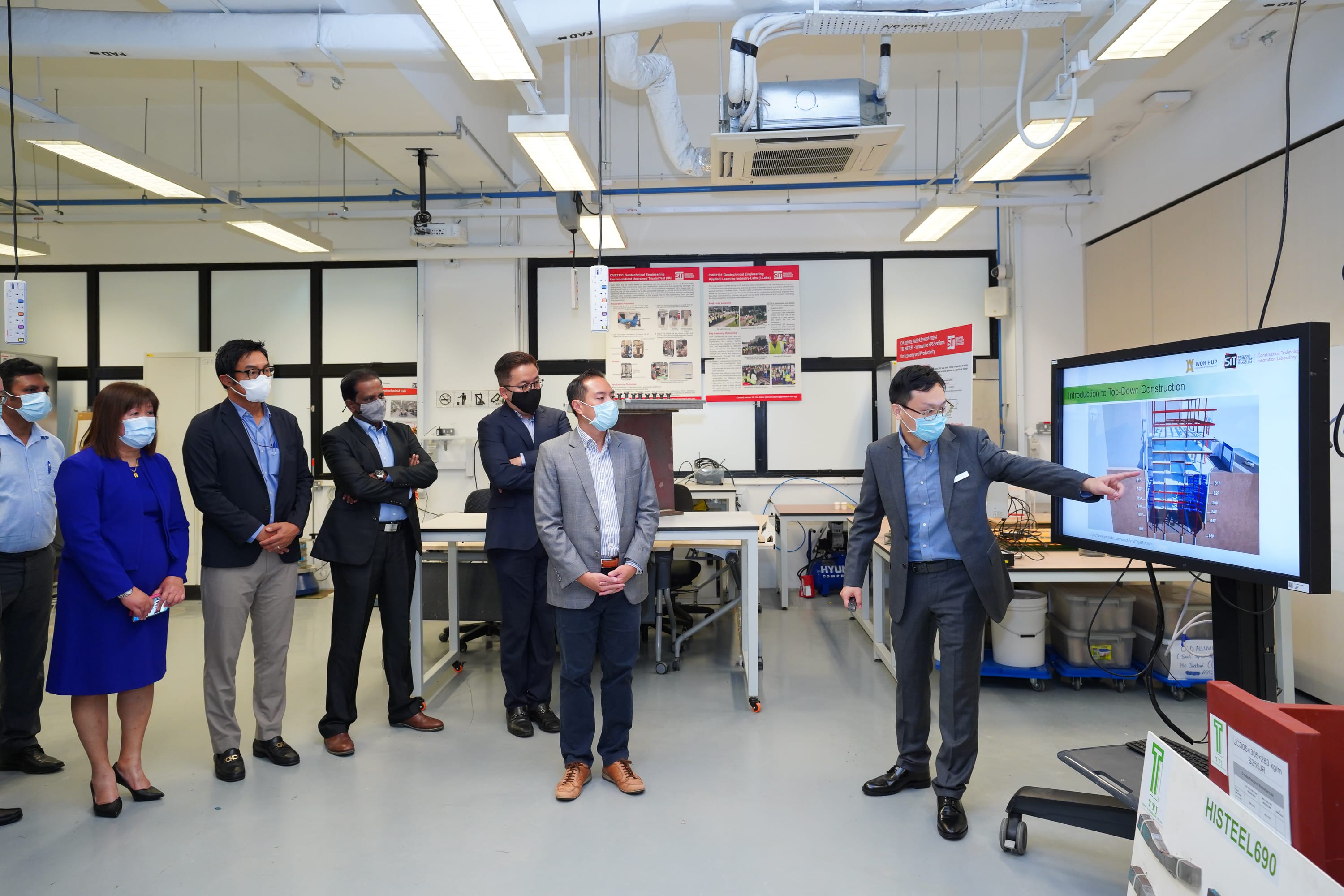 Group in masks watches presentation on "Top-Down Construction" with TTJ HISTEEL sign visible.