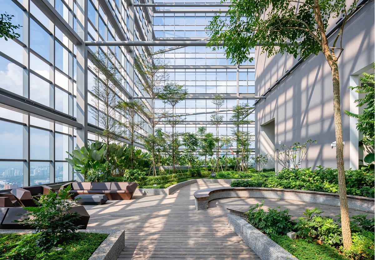 Rooftop garden with wooden walkways, benches, sofas, greenery, and a glass ceiling/wall.