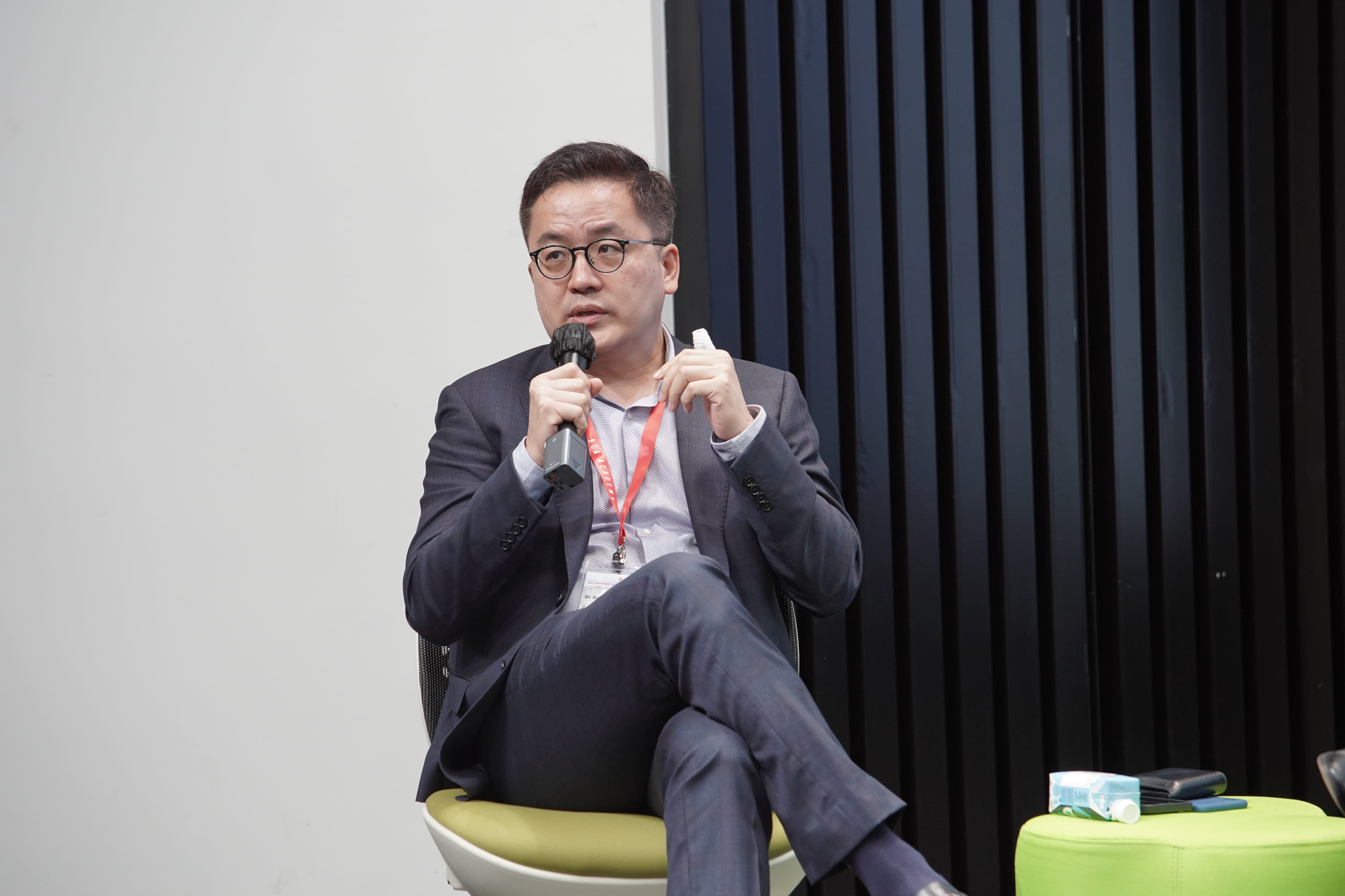 Man in a suit with glasses seated, holding a microphone, speaking.