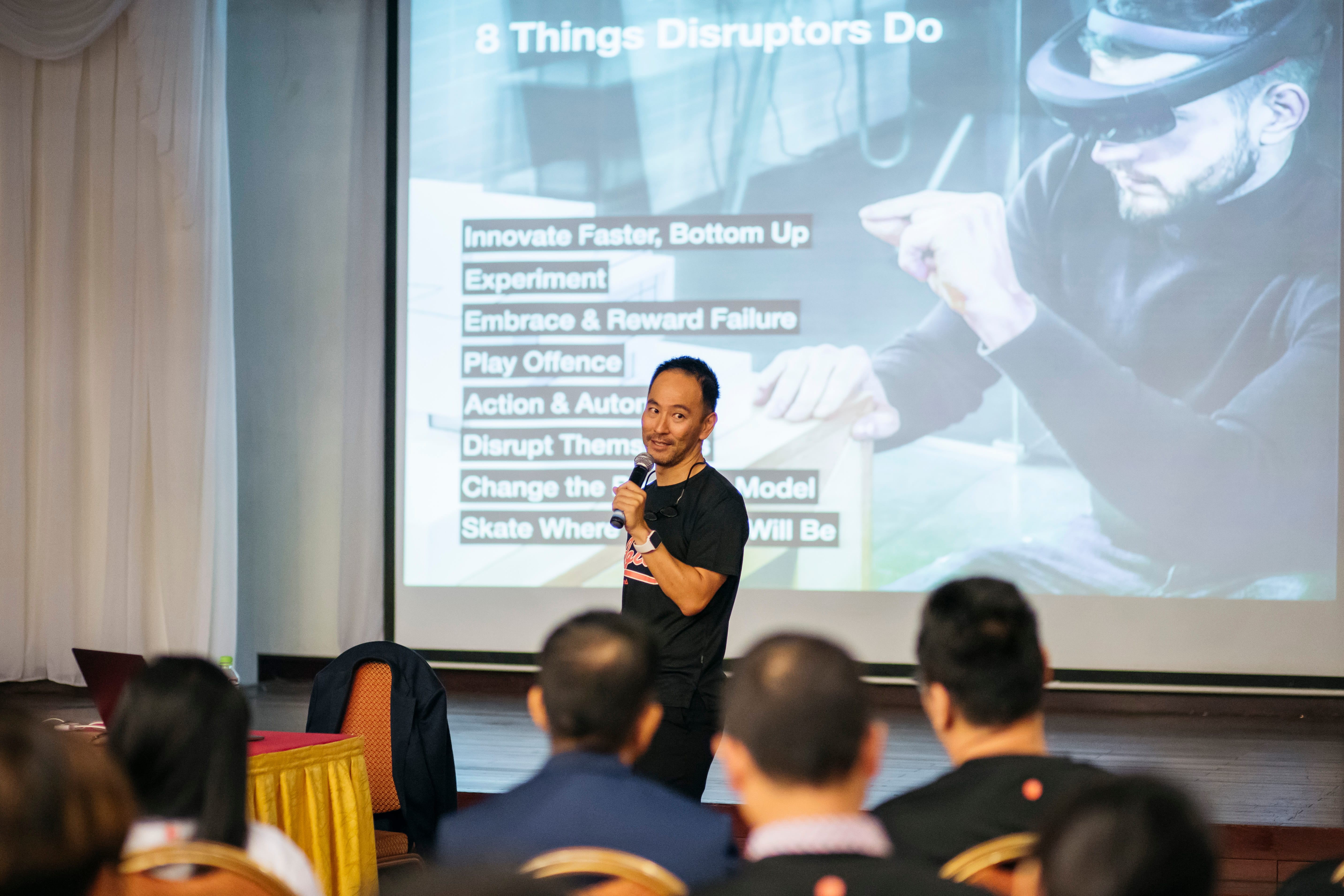 Speaker with mic presents "8 Things Disruptors Do" with a man in AR glasses projected behind him. Audience visible.