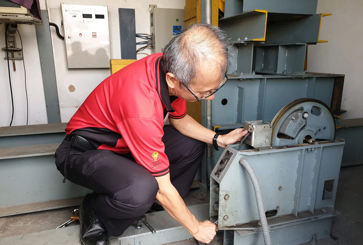Man in red shirt crouches, working on machinery with a large wheel and electrical boxes.