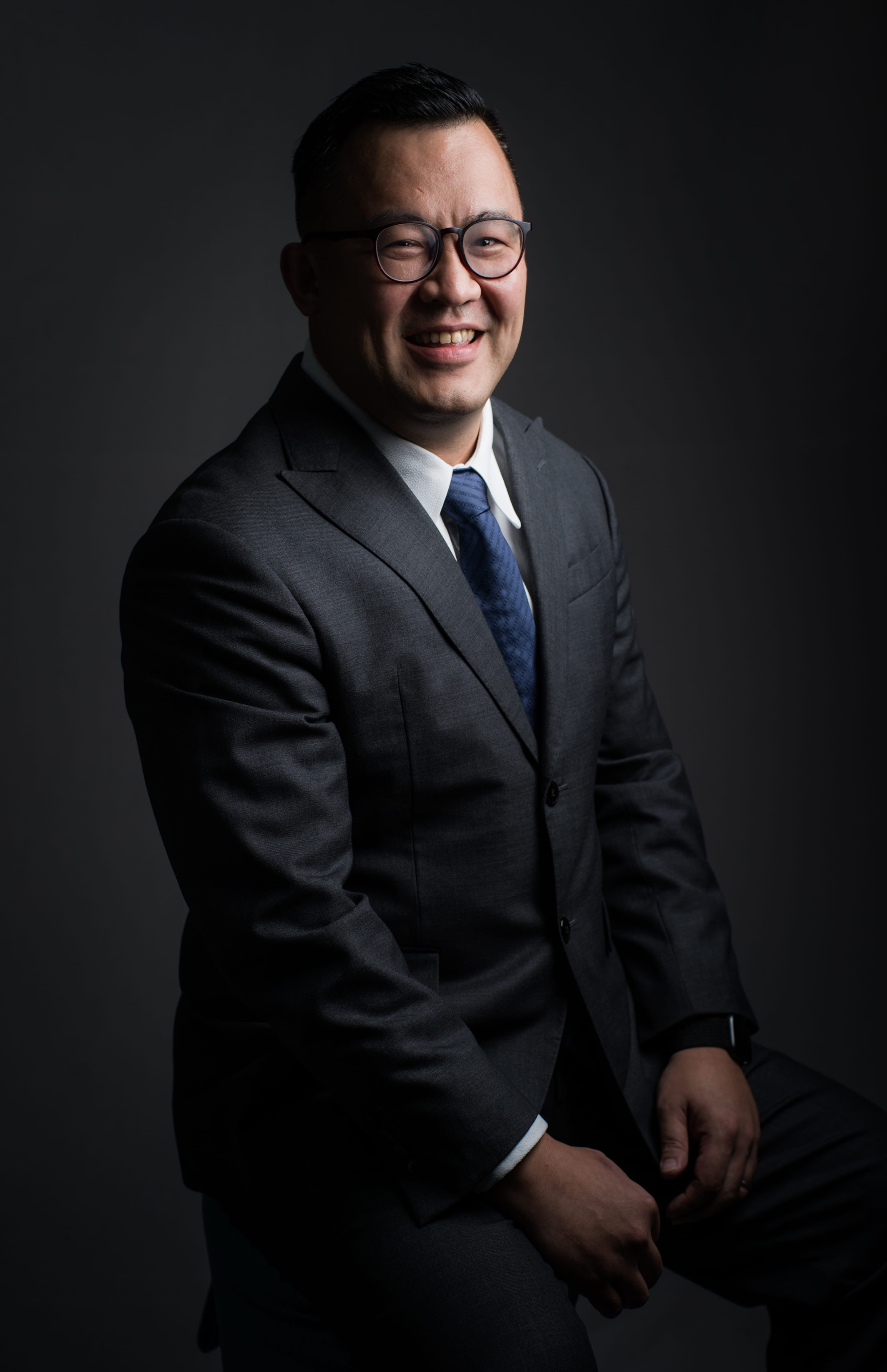 Smiling man in glasses, wearing a gray suit and blue tie, against a dark gray background.