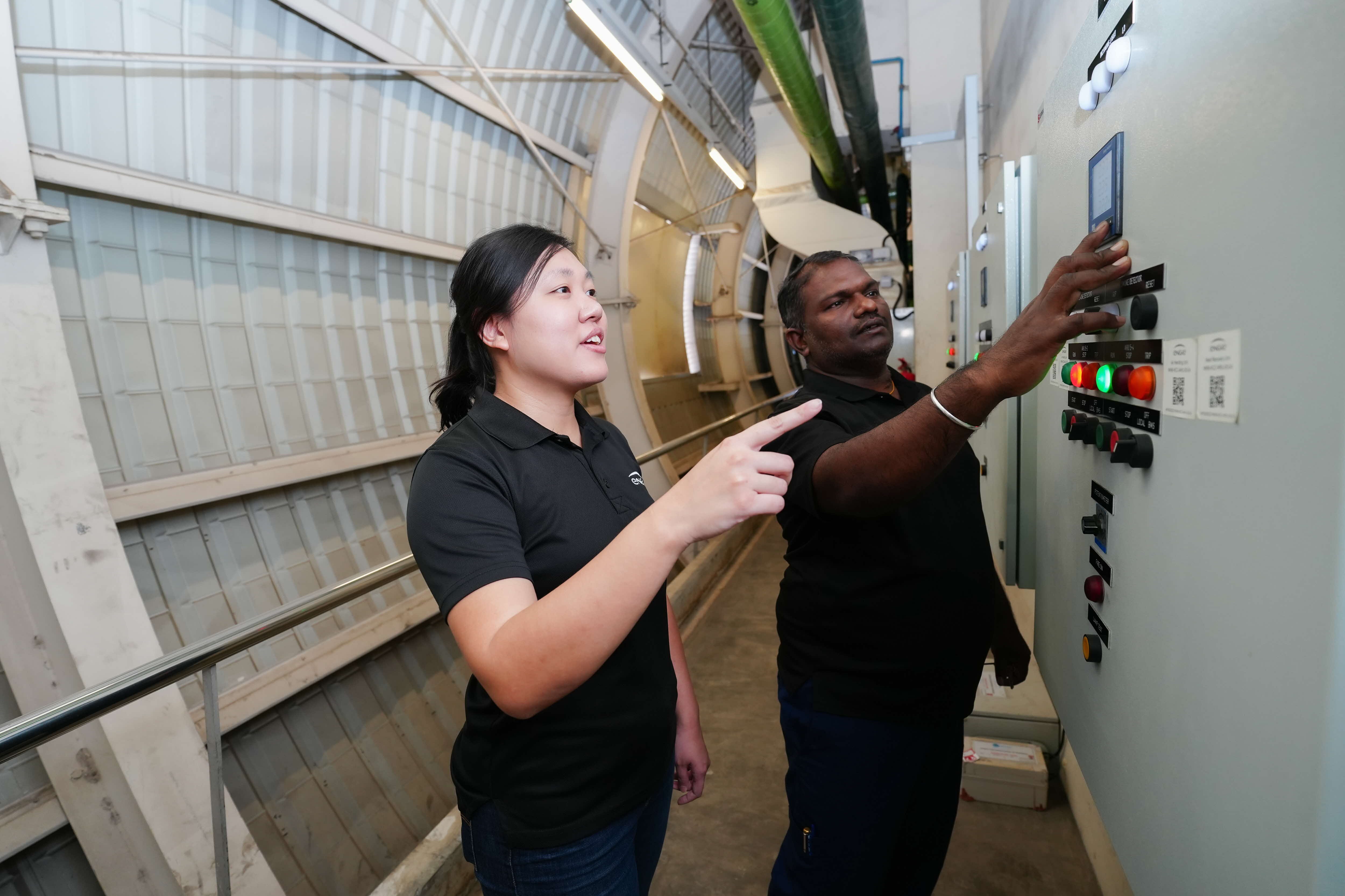 Two workers, one operating a control panel with lights and switches, in an industrial setting.
