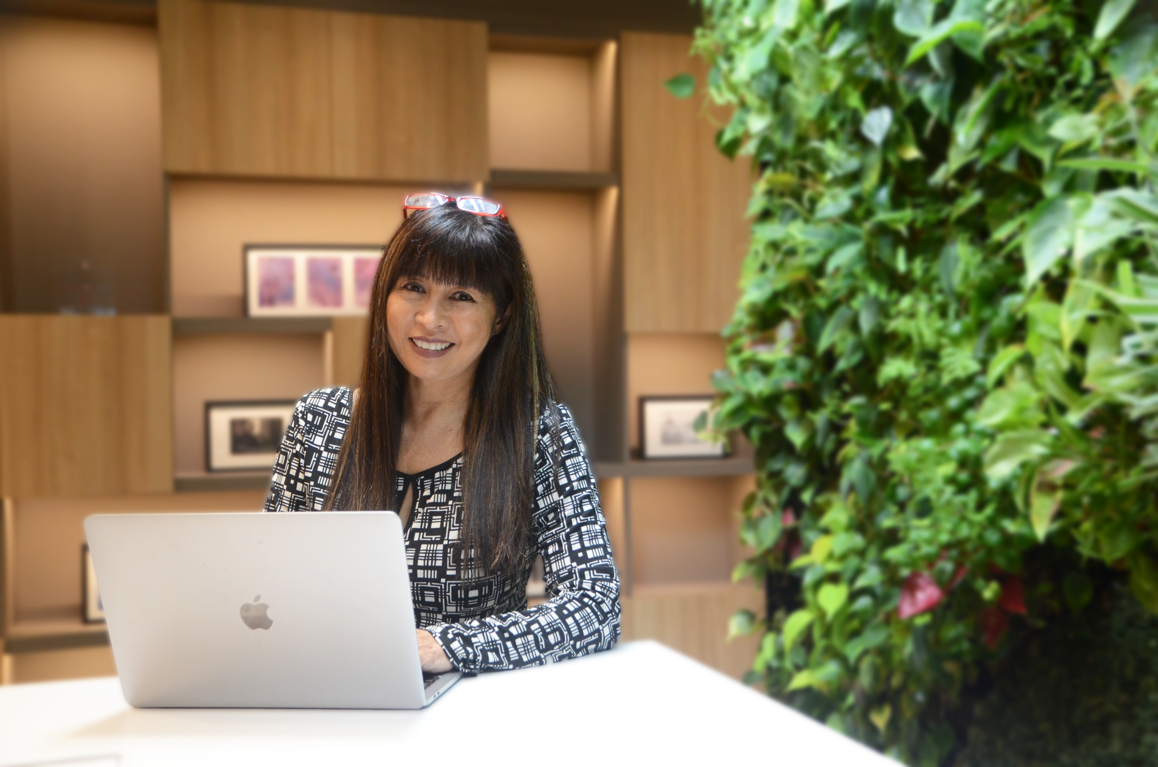 Woman with glasses using a MacBook in a room with a green plant wall.