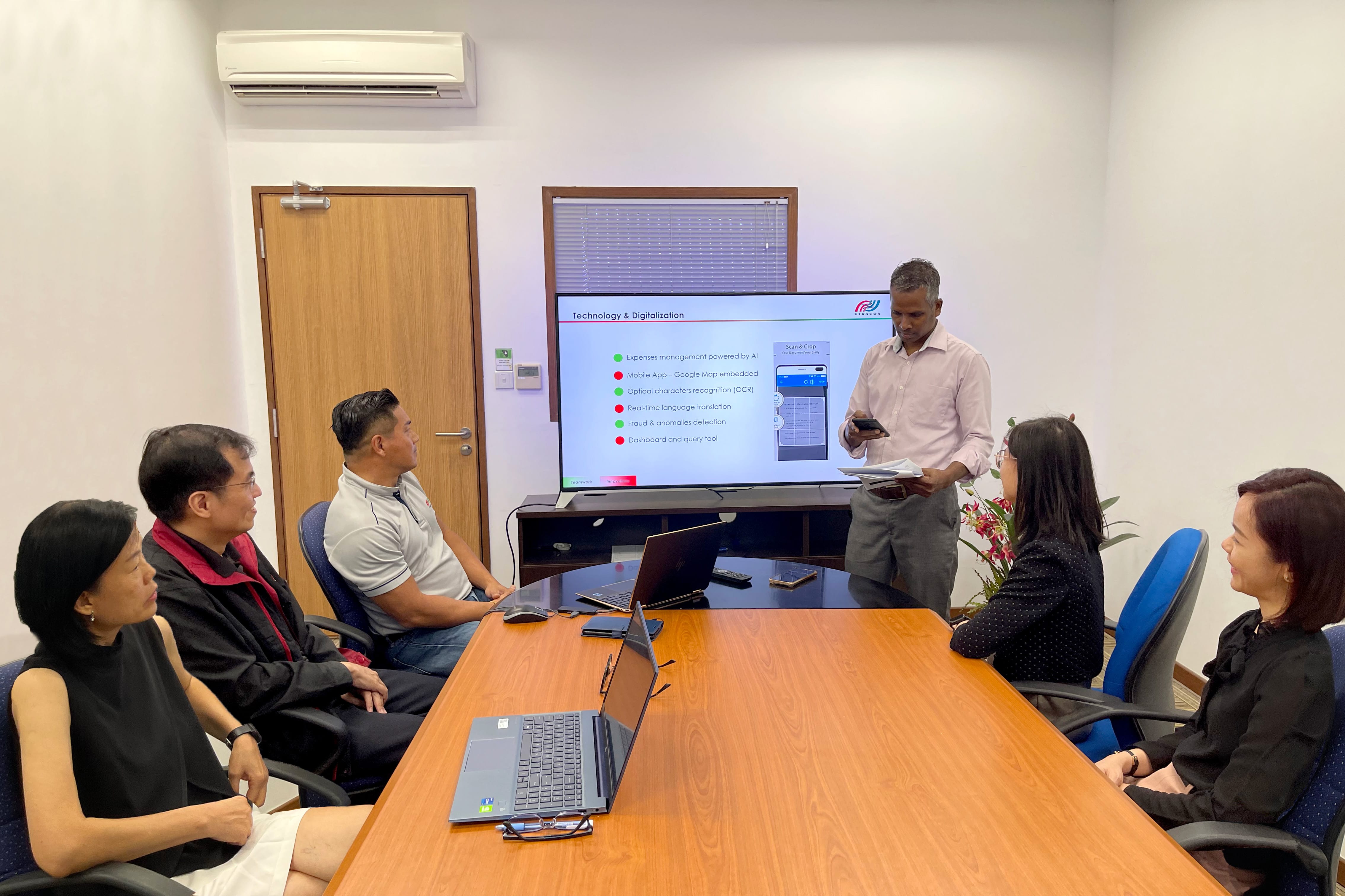 People in a conference room, man presenting "Technology & Digitalization" on a large display.
