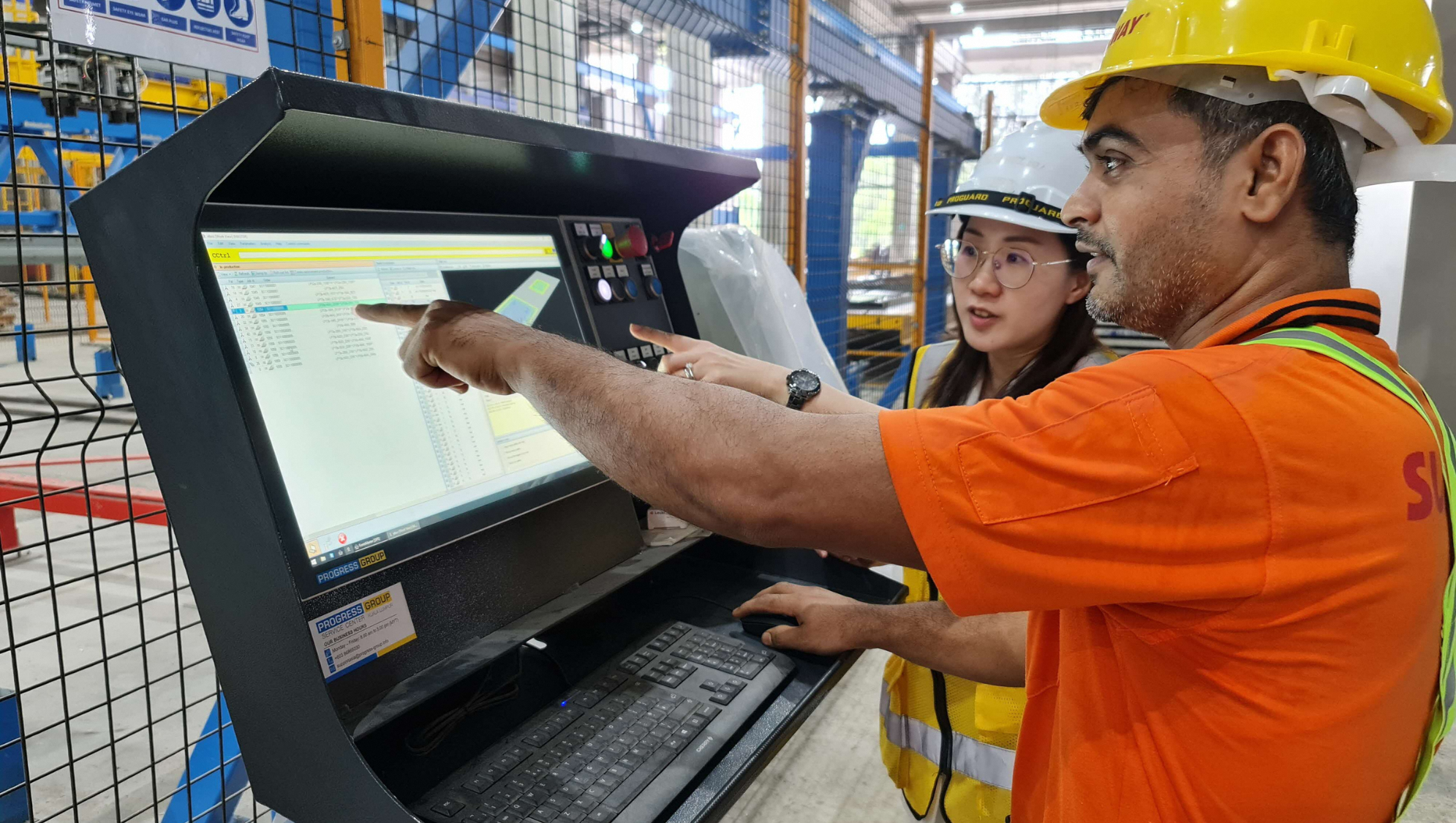Two workers point at a computer screen on a manufacturing control console.