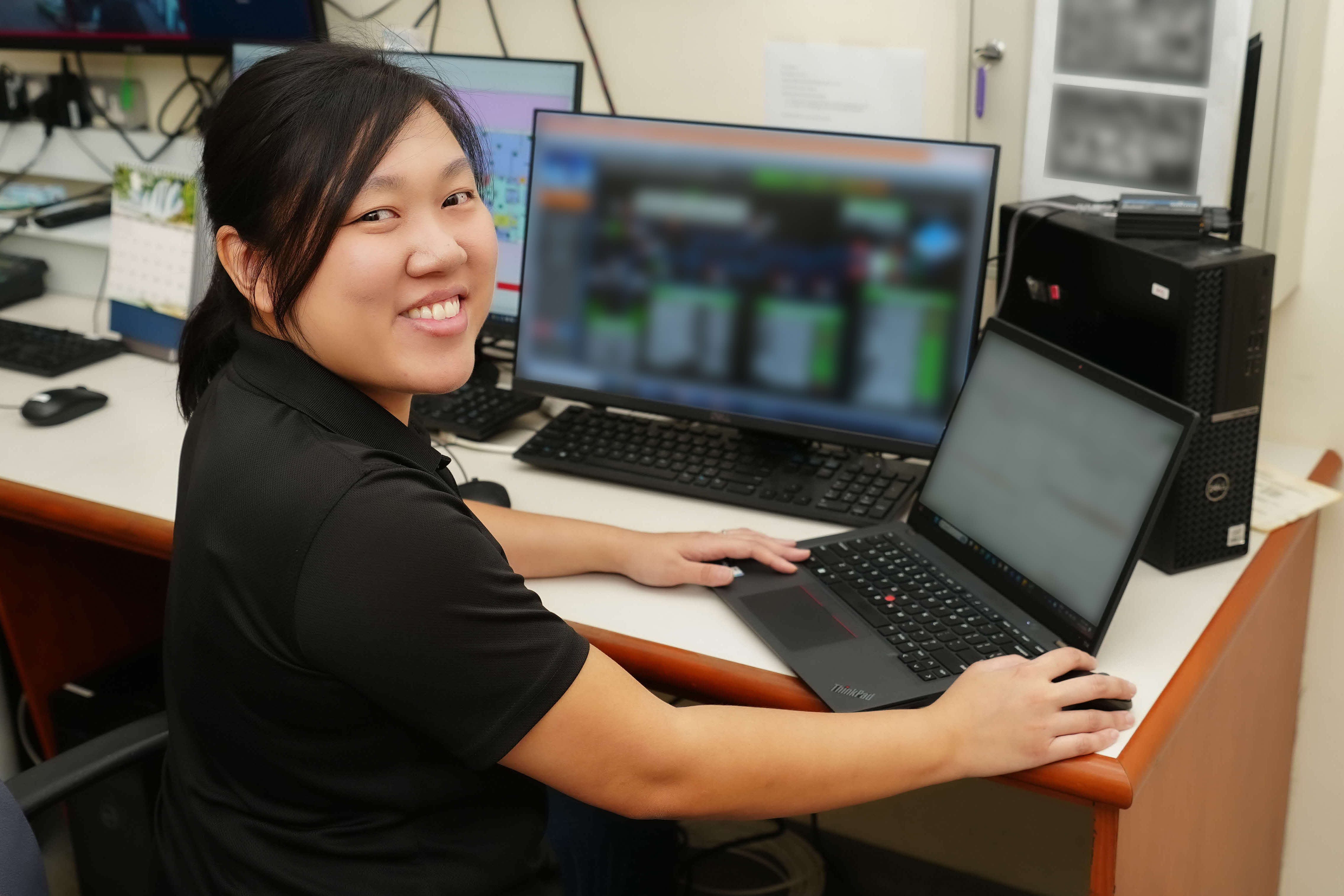 Smiling woman works at a desk with monitors, a Dell PC, and a ThinkPad laptop.