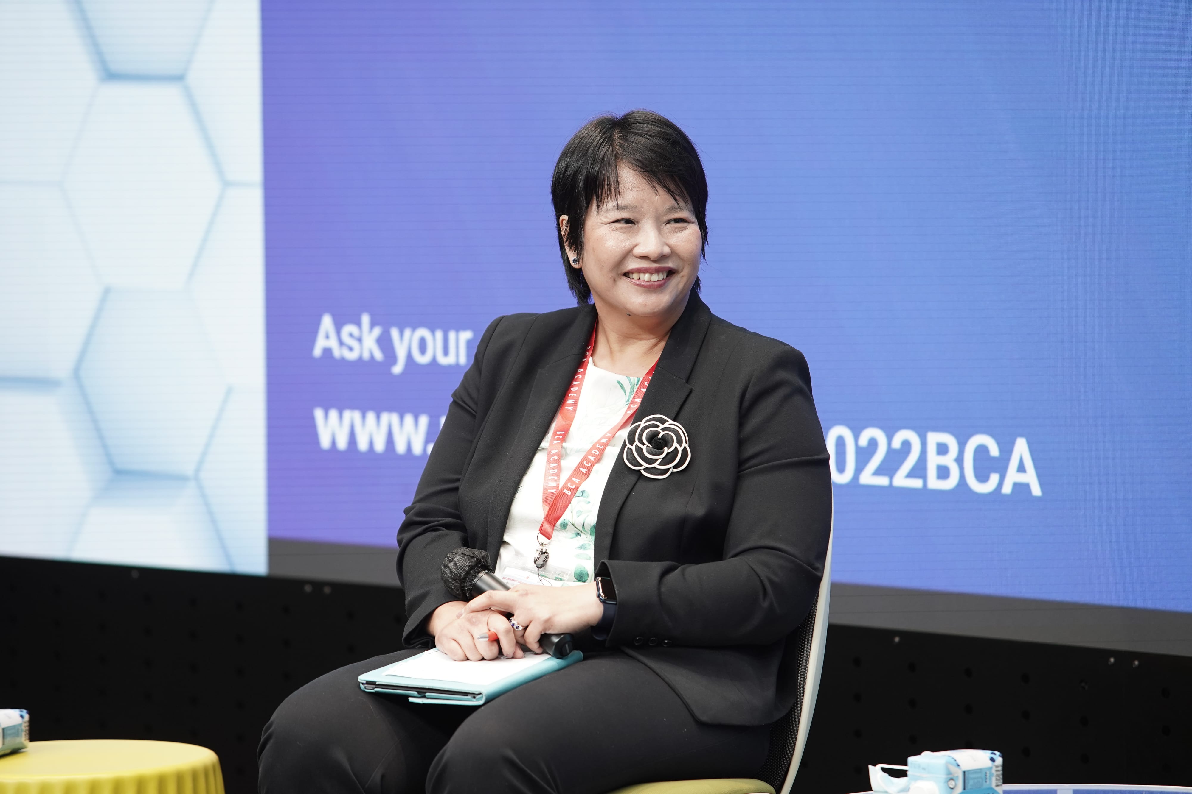 Woman in black suit with BCA Academy lanyard, seated, holding mic and clipboard, smiles. Blue screen behind.