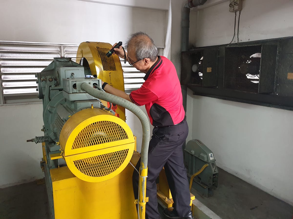 Man in red shirt inspecting industrial equipment with flashlight. Yellow safety guards visible.