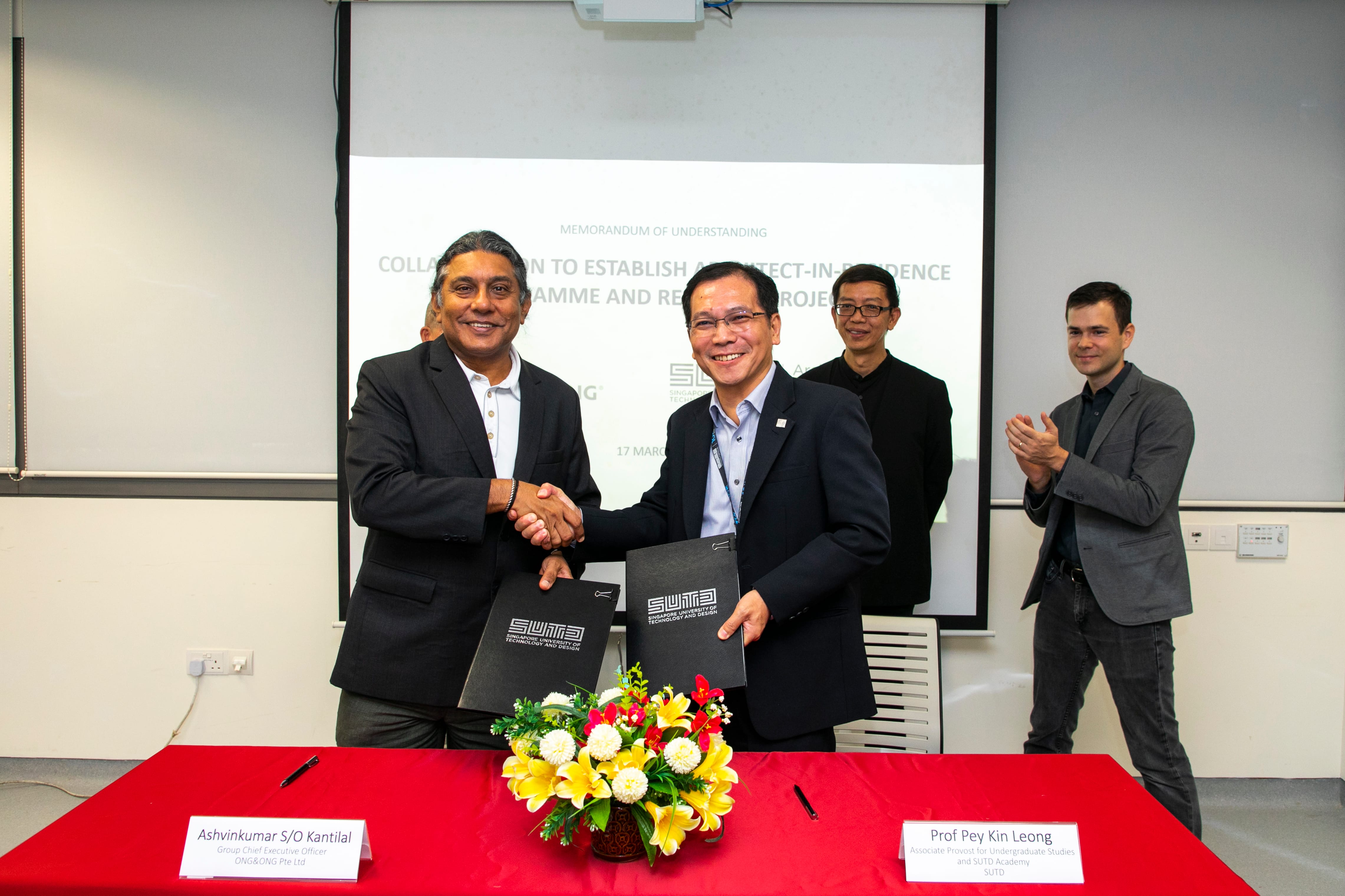Two men in suits shaking hands, holding SUTD documents, behind them two men and a screen.