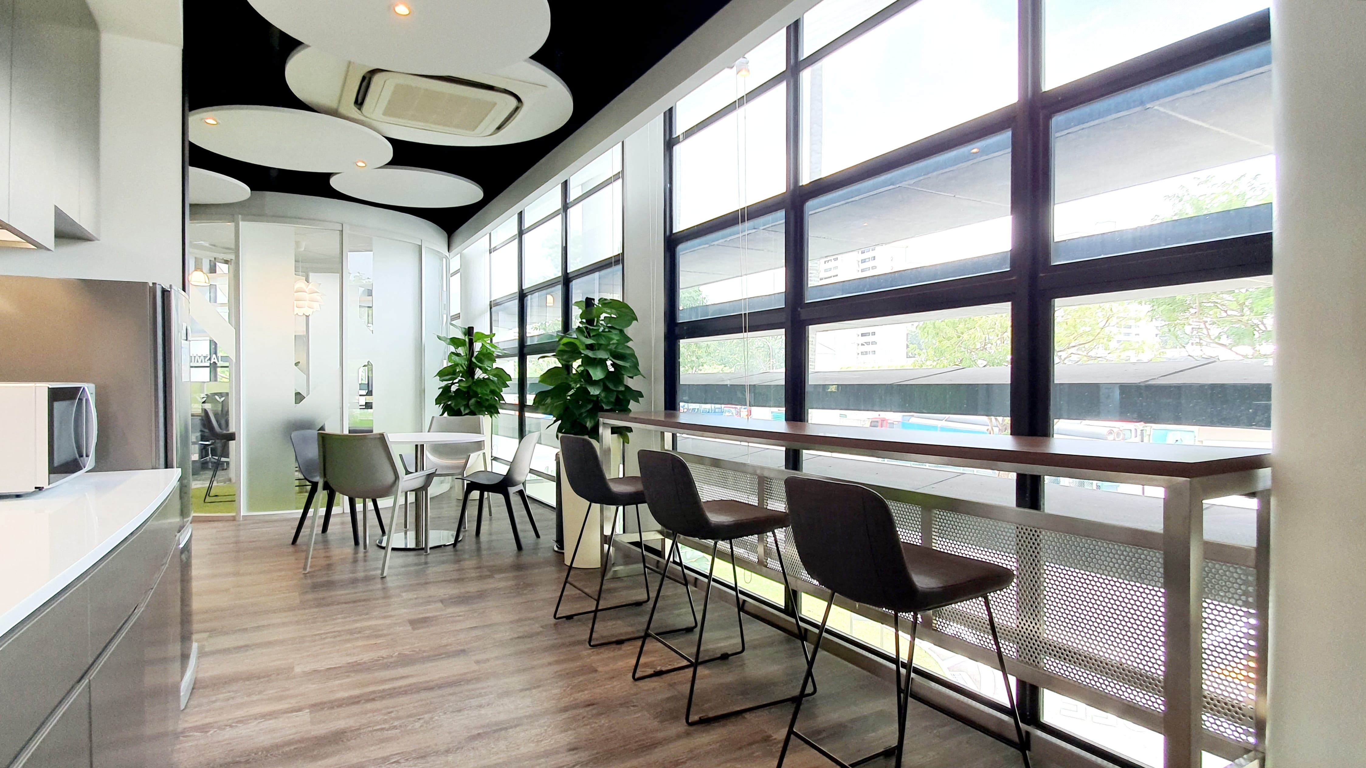 Modern office space with bar stools by large windows, potted plants, and kitchen area.