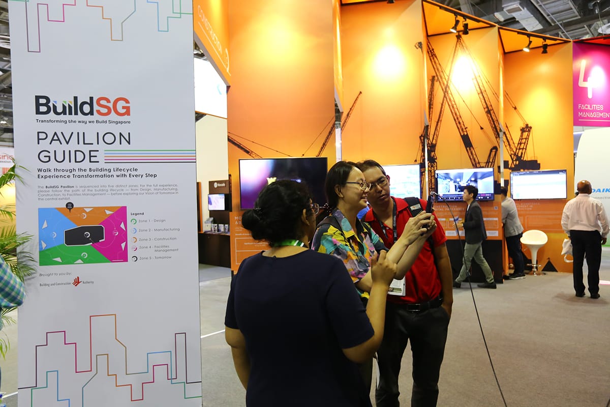 Attendees at BuildSG Pavilion with crane backdrop, one taking photos with phone.