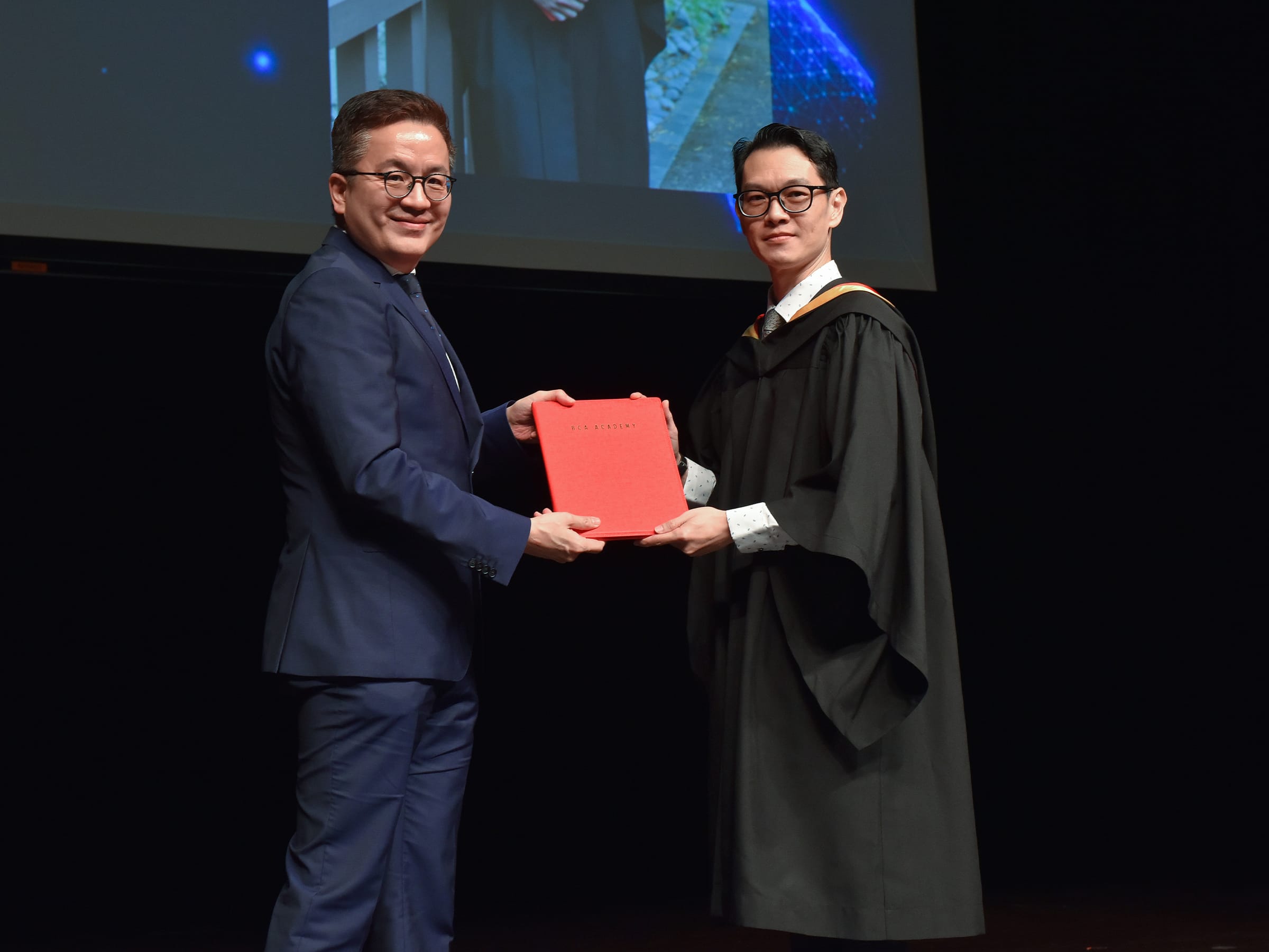 Two men exchange red "BCA Academy" document. One in suit, one in graduation gown.
