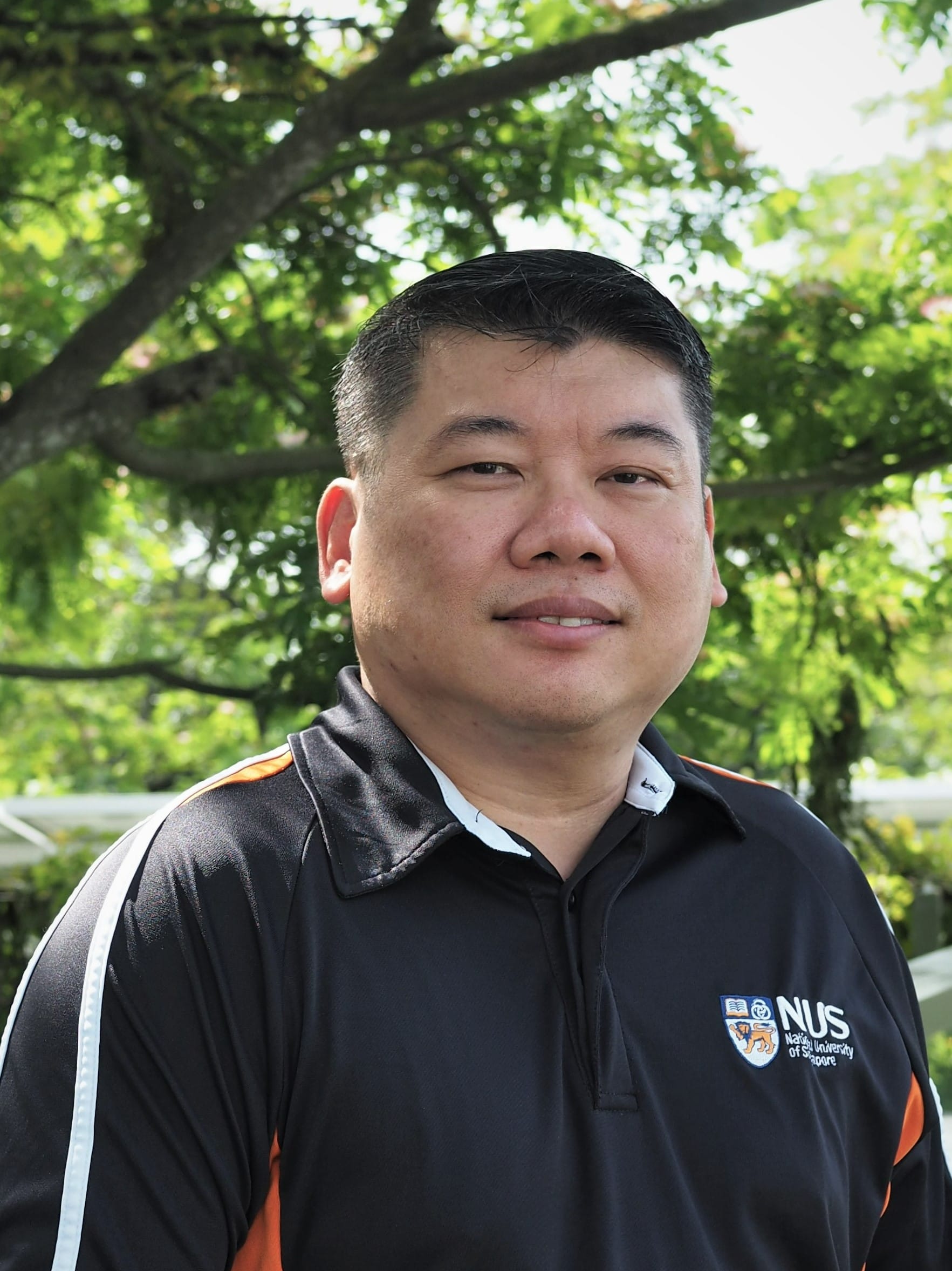 Man in black NUS polo shirt with orange/white trim, blurred green background.