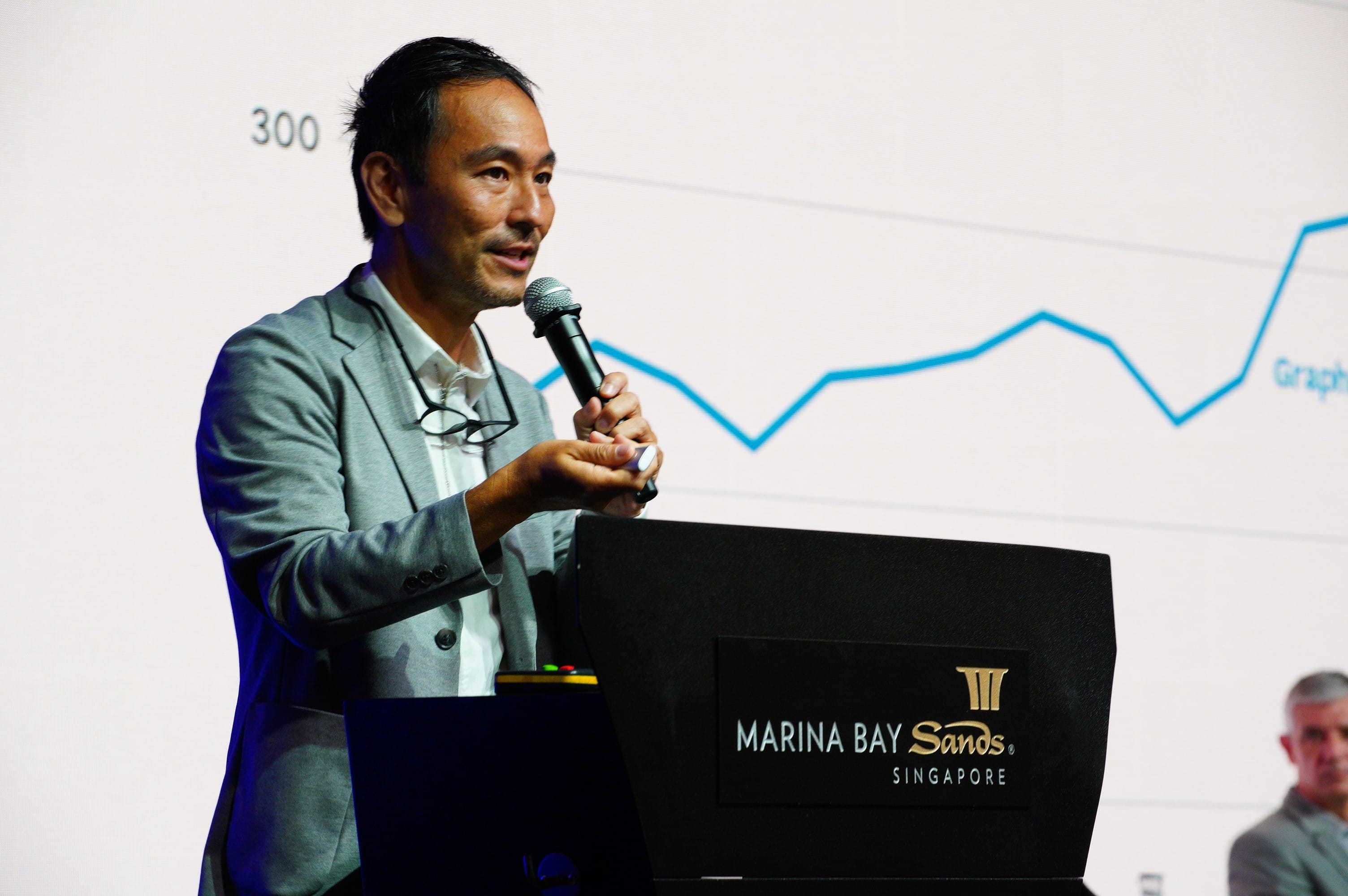 Man at Marina Bay Sands podium speaks into mic, glasses hanging. Graph background.
