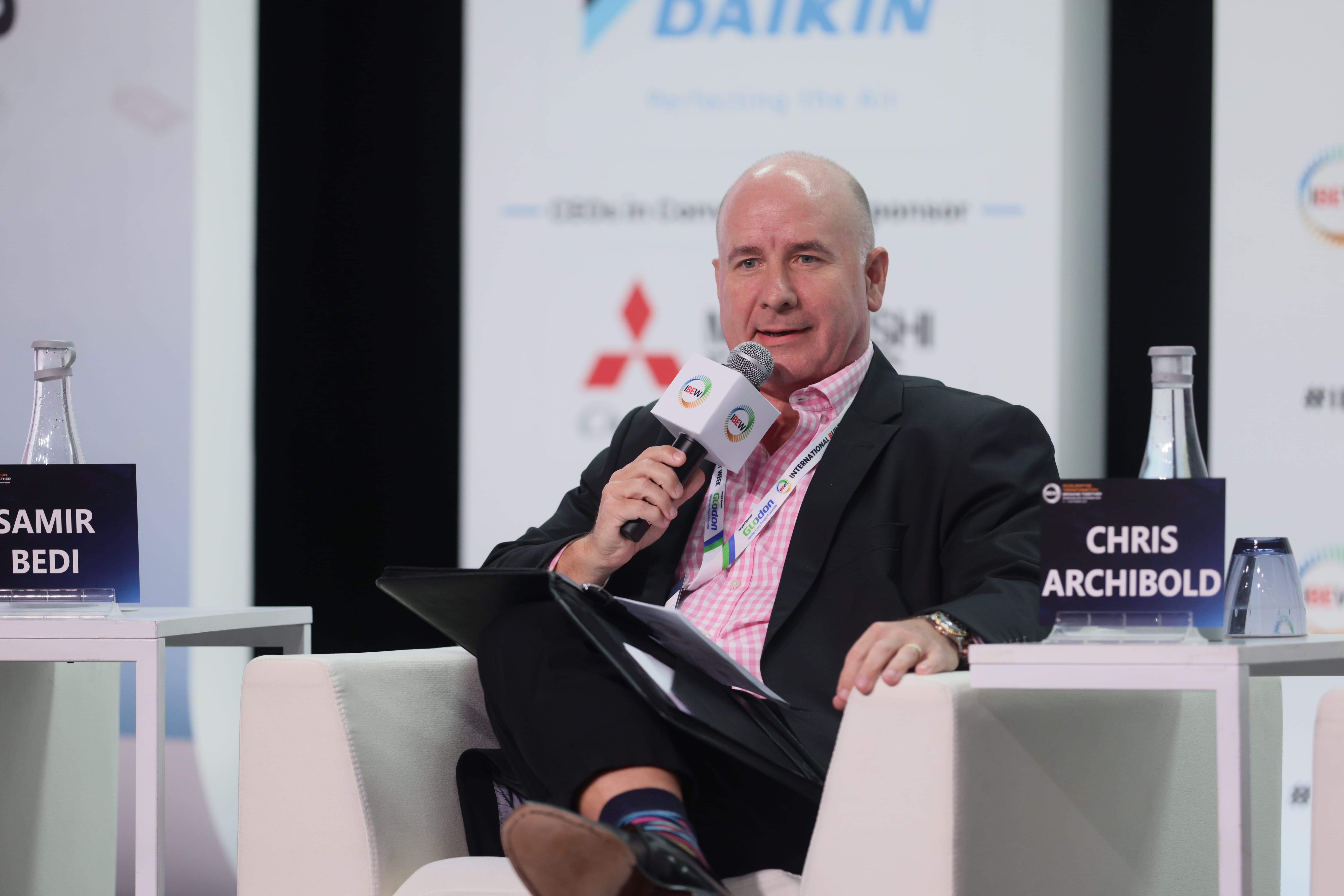 Man in blazer holding IBEW mic on panel; Daikin and Mitsubishi logos, nameplates read "Samir Bedi" and "Chris Archibold."