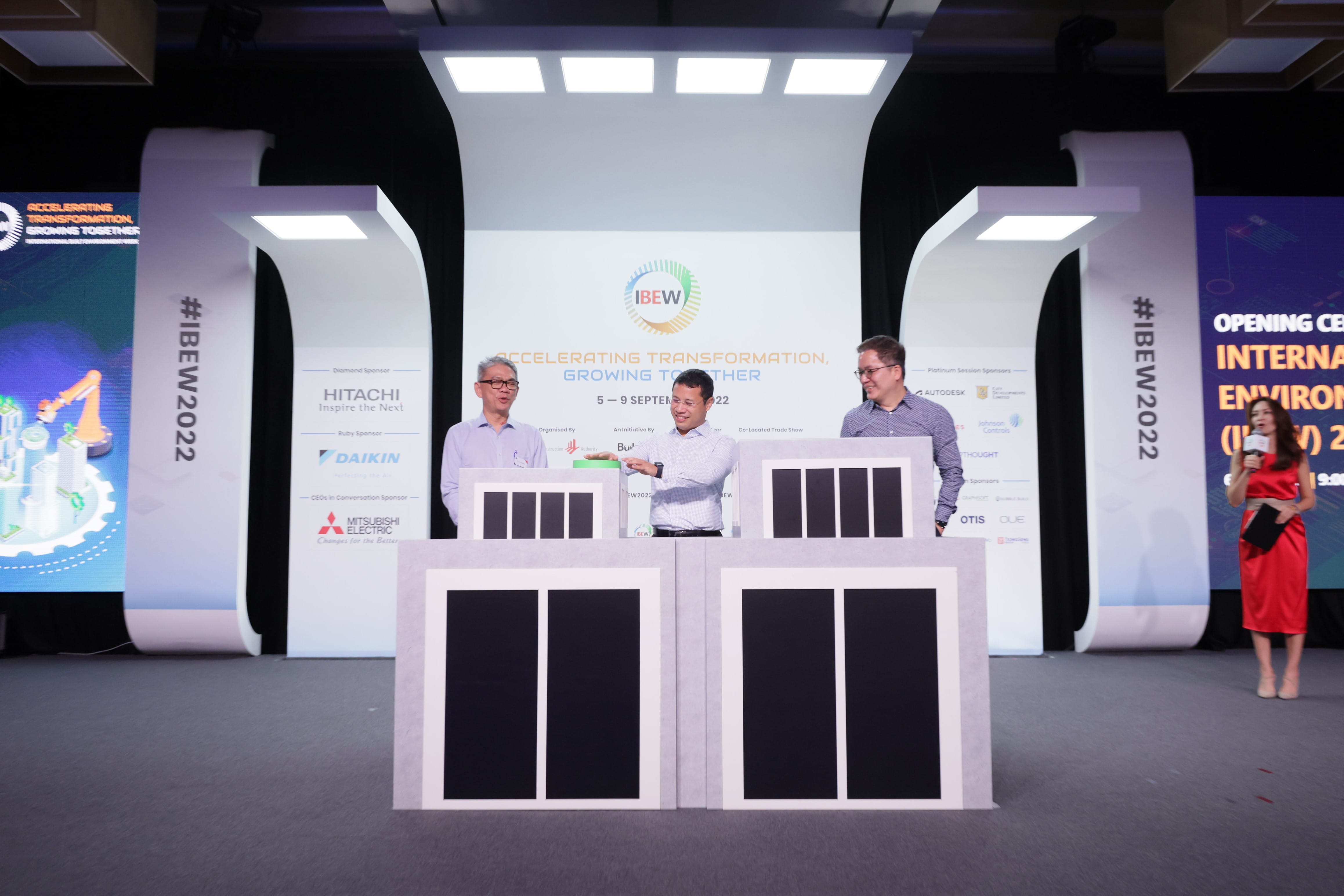 Three people on stage at IBEW event with Hitachi, Daikin and Mitsubishi Electric sponsor logos. Woman with microphone.