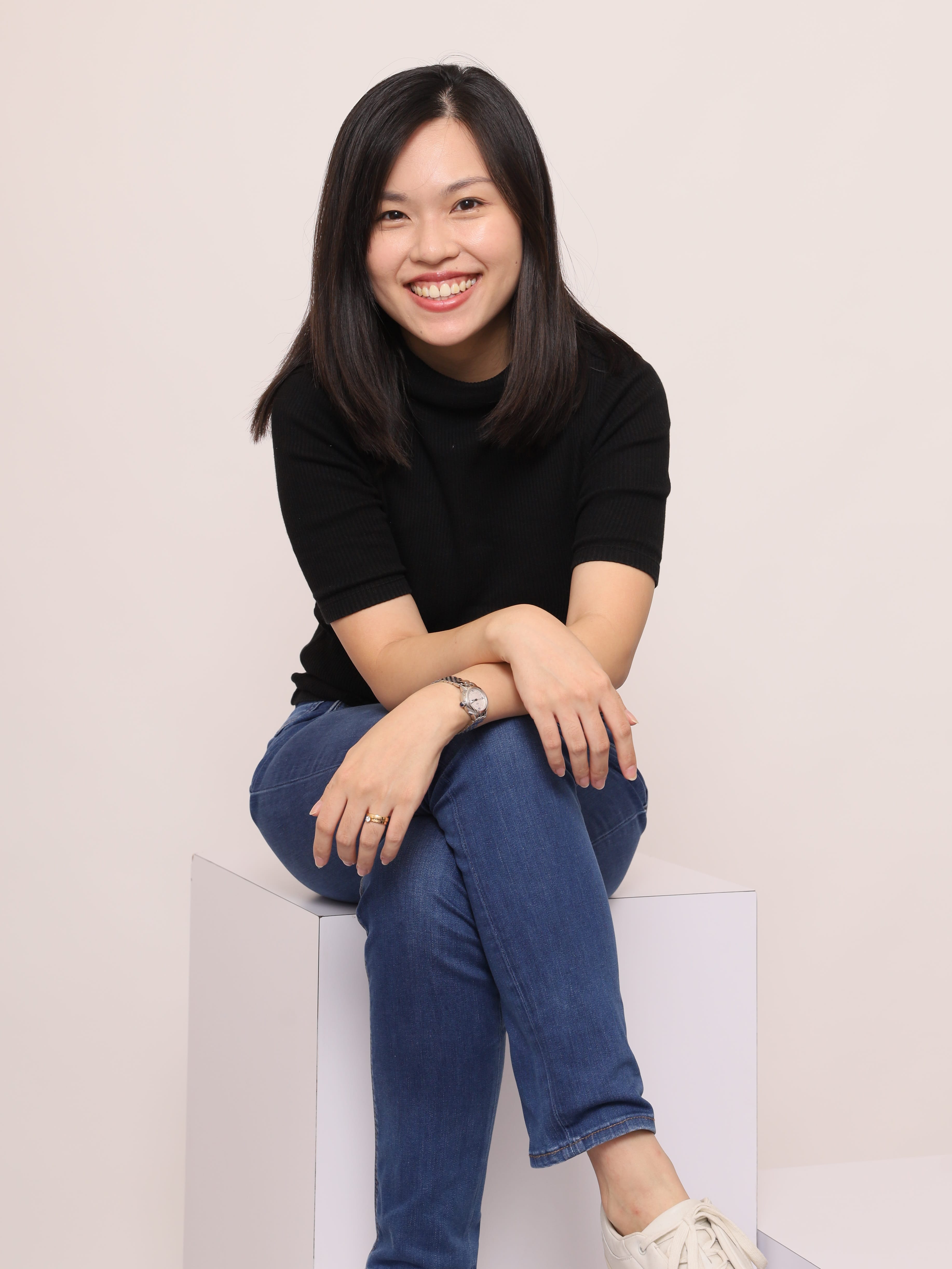 Woman with long dark hair, wearing blue jeans and a black top, seated on a white block.