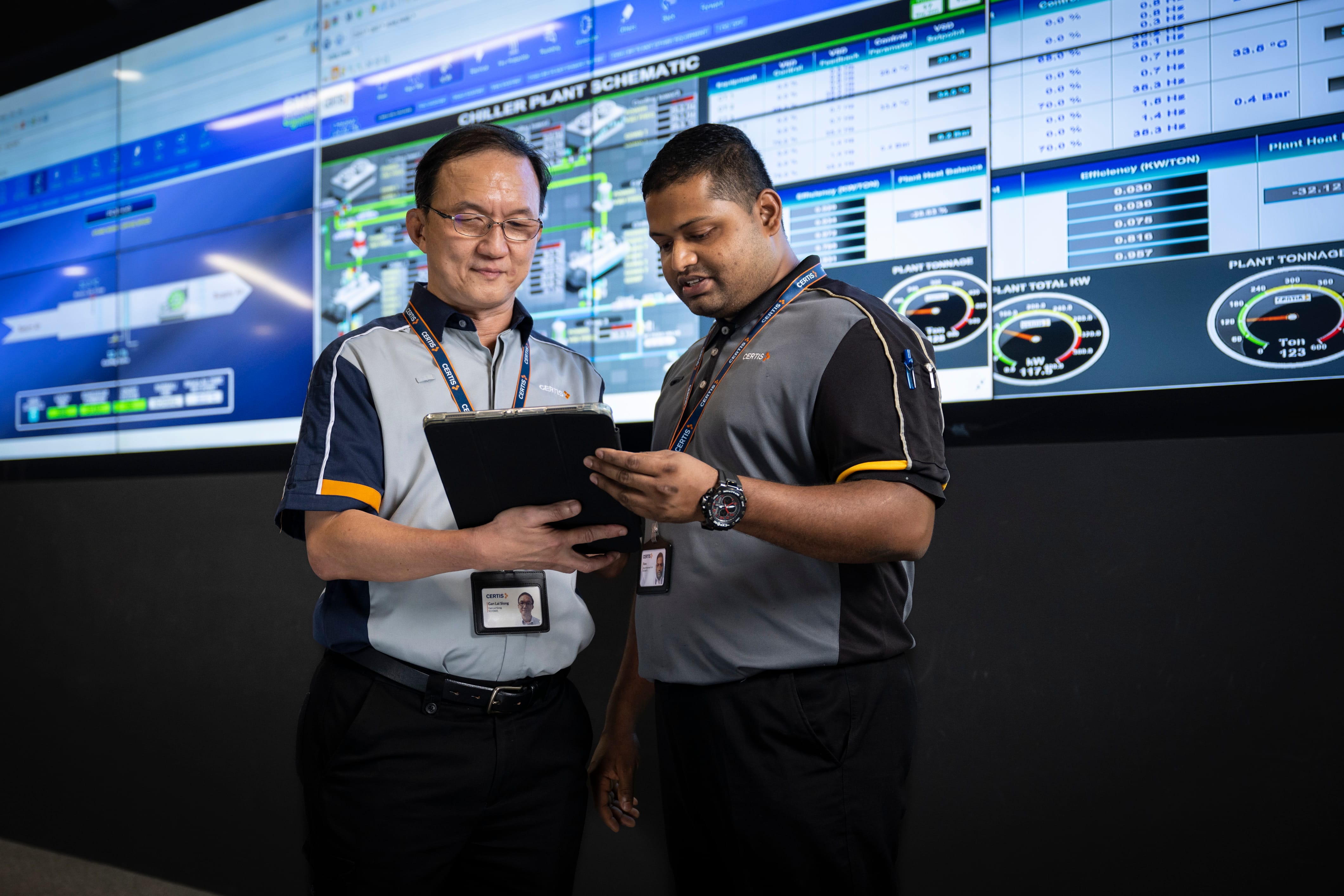 Two Certis employees in uniform reviewing data on a tablet in front of a large chiller plant schematic display.