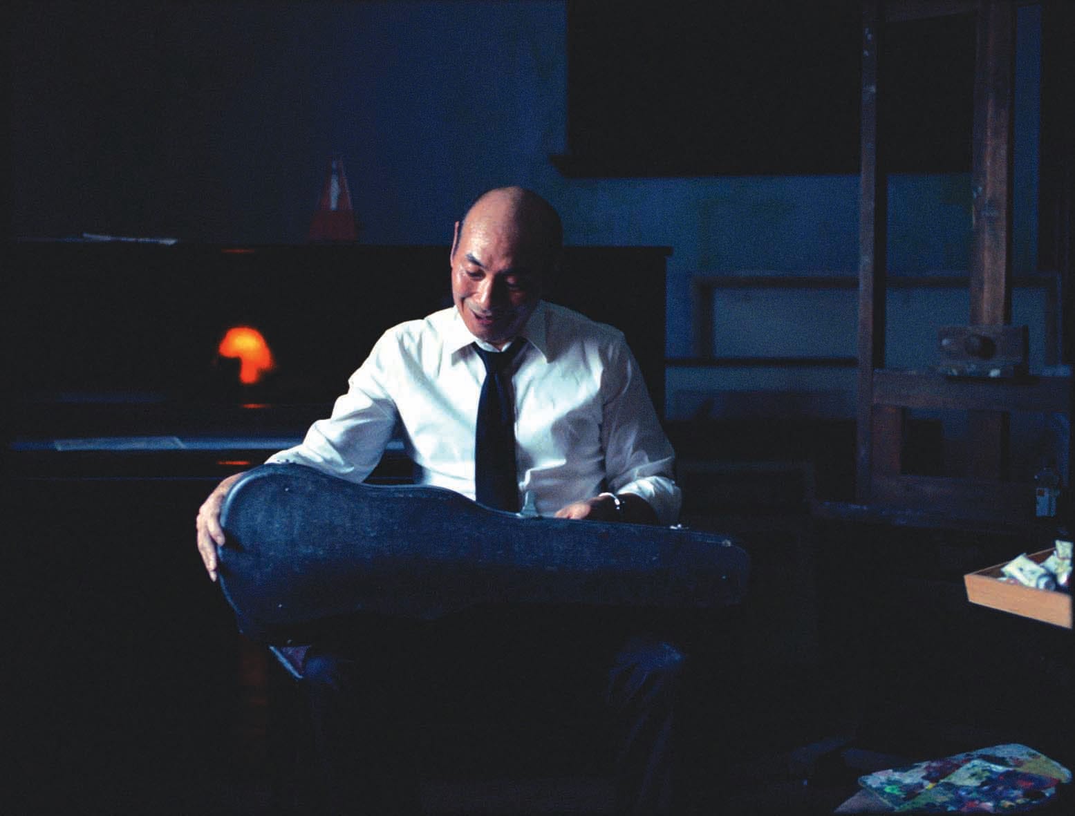 A man in a white shirt and black tie sits in a dimly lit room holding a guitar case.