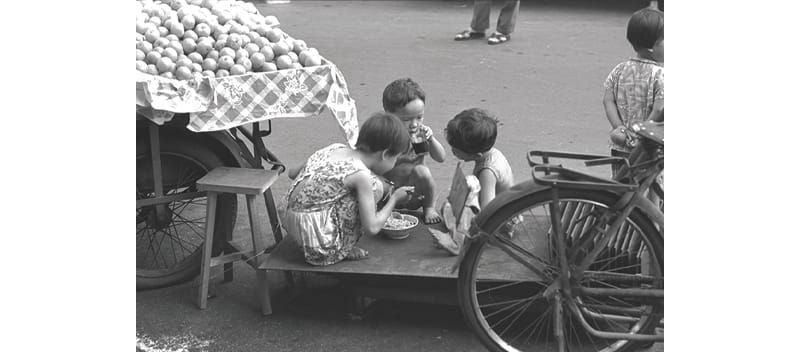 Three children sit on a wooden platform, eating next to a bicycle and a cart filled with fruit covered by a cloth.