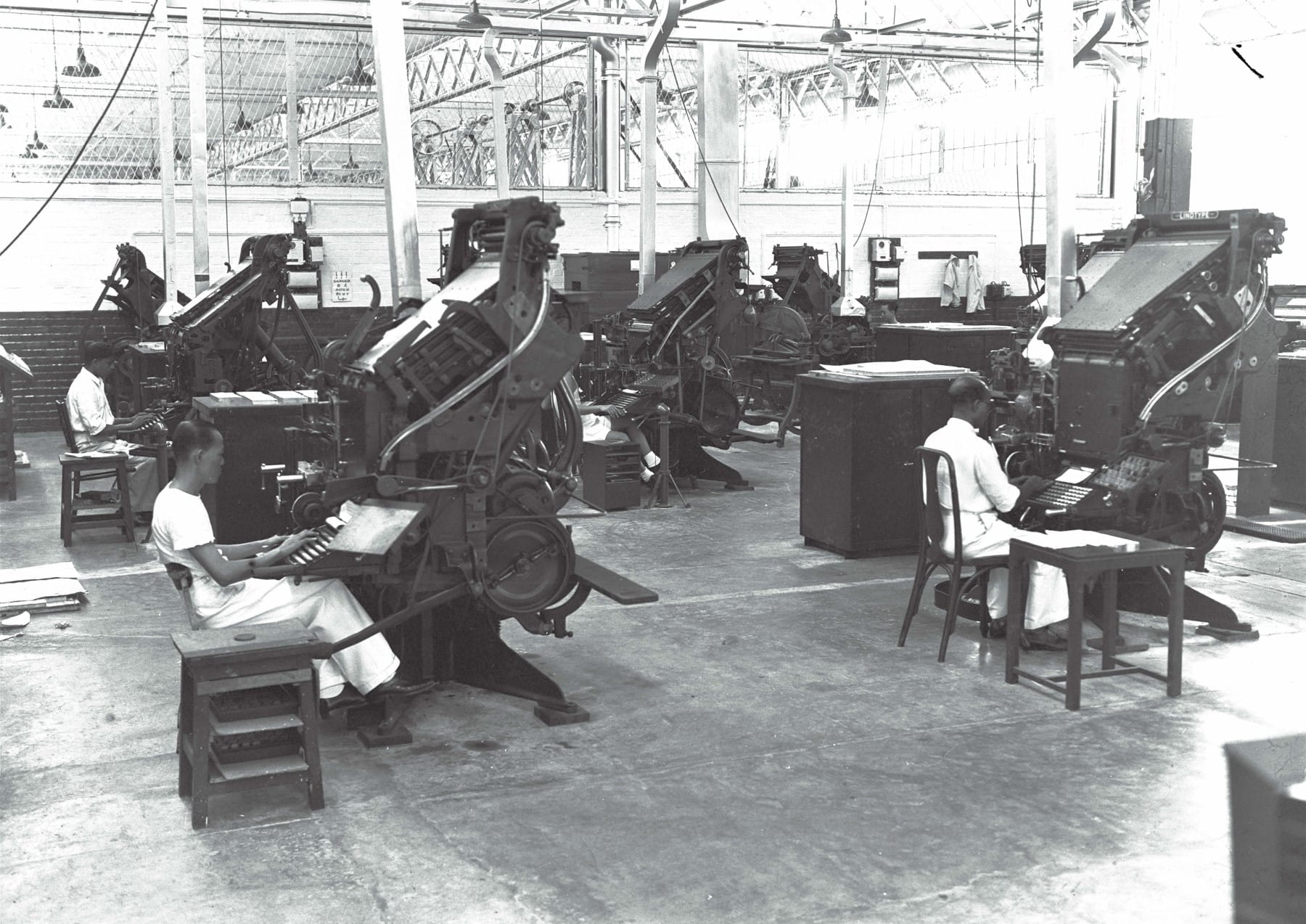 People are operating large printing machines in a spacious, industrial workshop with high ceilings and overhead lighting.