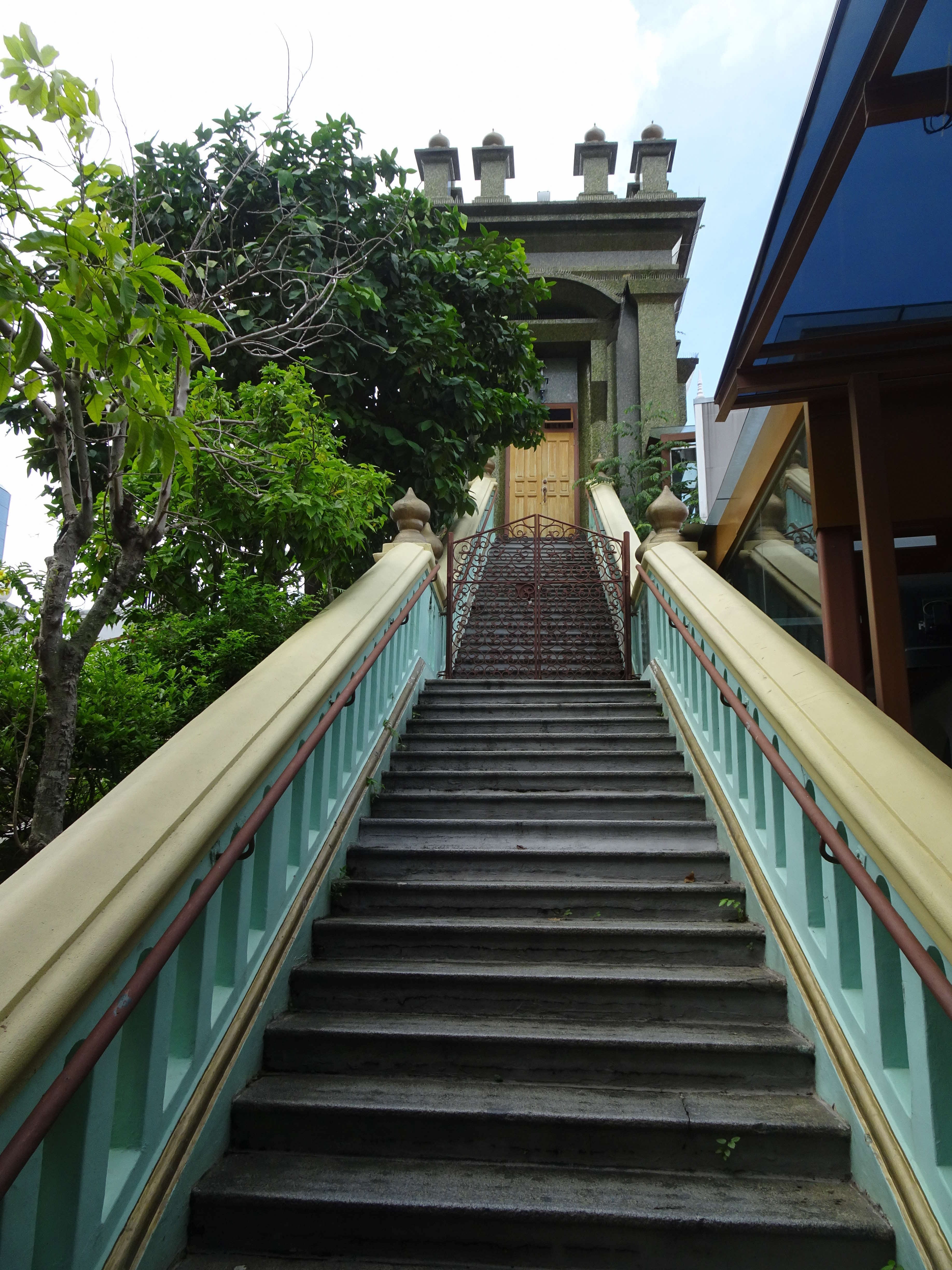 Concrete stairs with beige and teal railings leading up to a building with wooden doors, with trees on either side.