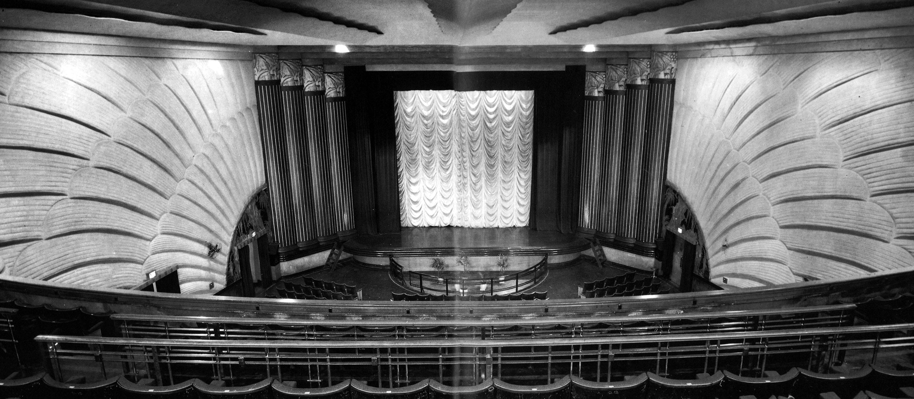 Black and white image of an empty, ornate theater with curved walls and a stage featuring a curtain.