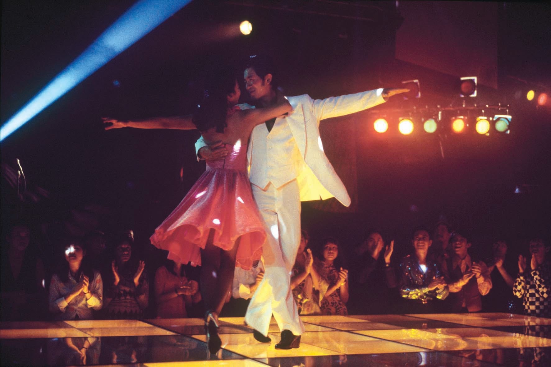 A couple dances under colorful lights on a disco floor while an audience claps and watches them.