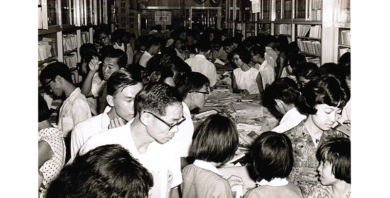 A crowded library with people reading and browsing books on tables and shelves.