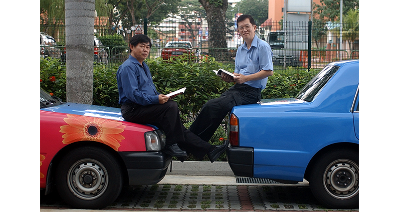 Two men sit on a red and a blue car, reading papers, with greenery and buildings in the background.