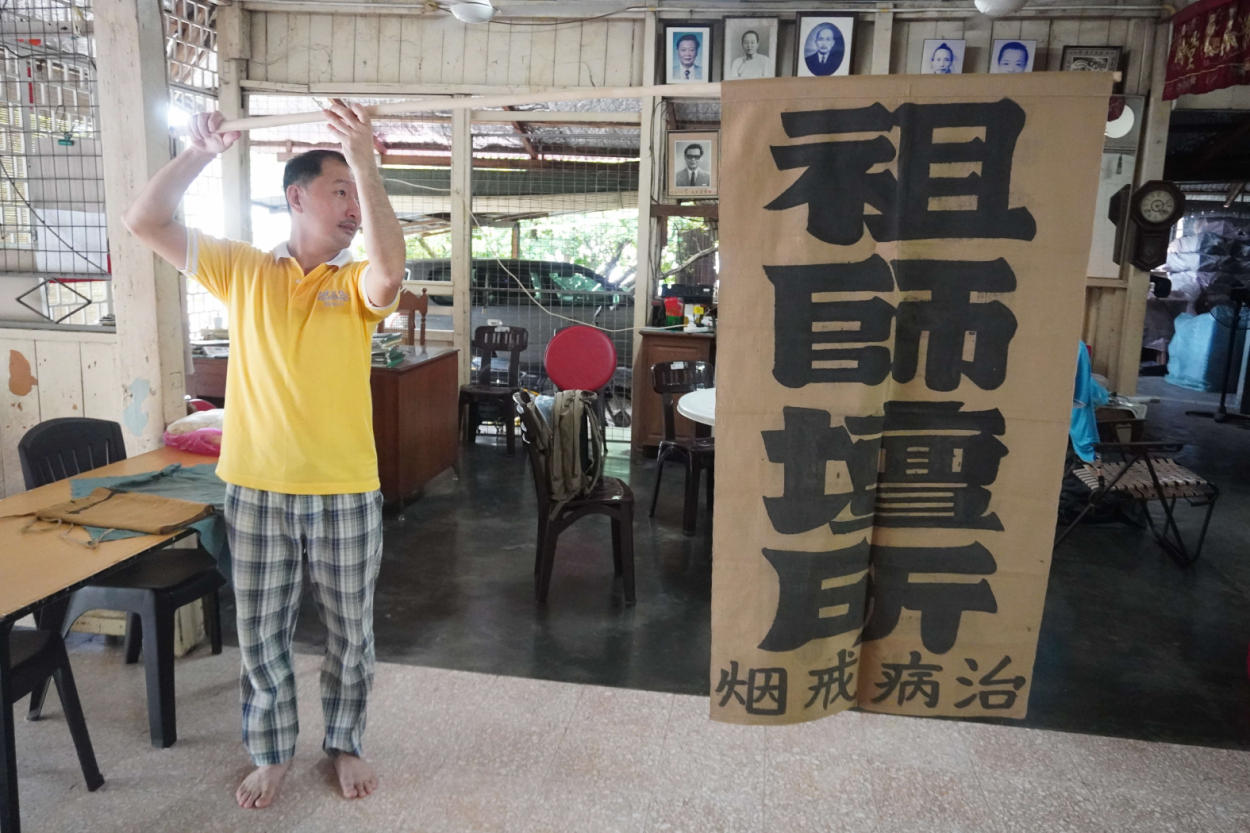 A banner owned by Huang Shengfa advertising Fook Poon Yuen Temple’s service of treating opium addicts and curing illnesses. It was hung from the temple’s second floor when it operated out of a shophouse. The banner is held by Huang’s grandson, 2019. Photograph by Esmond Soh.